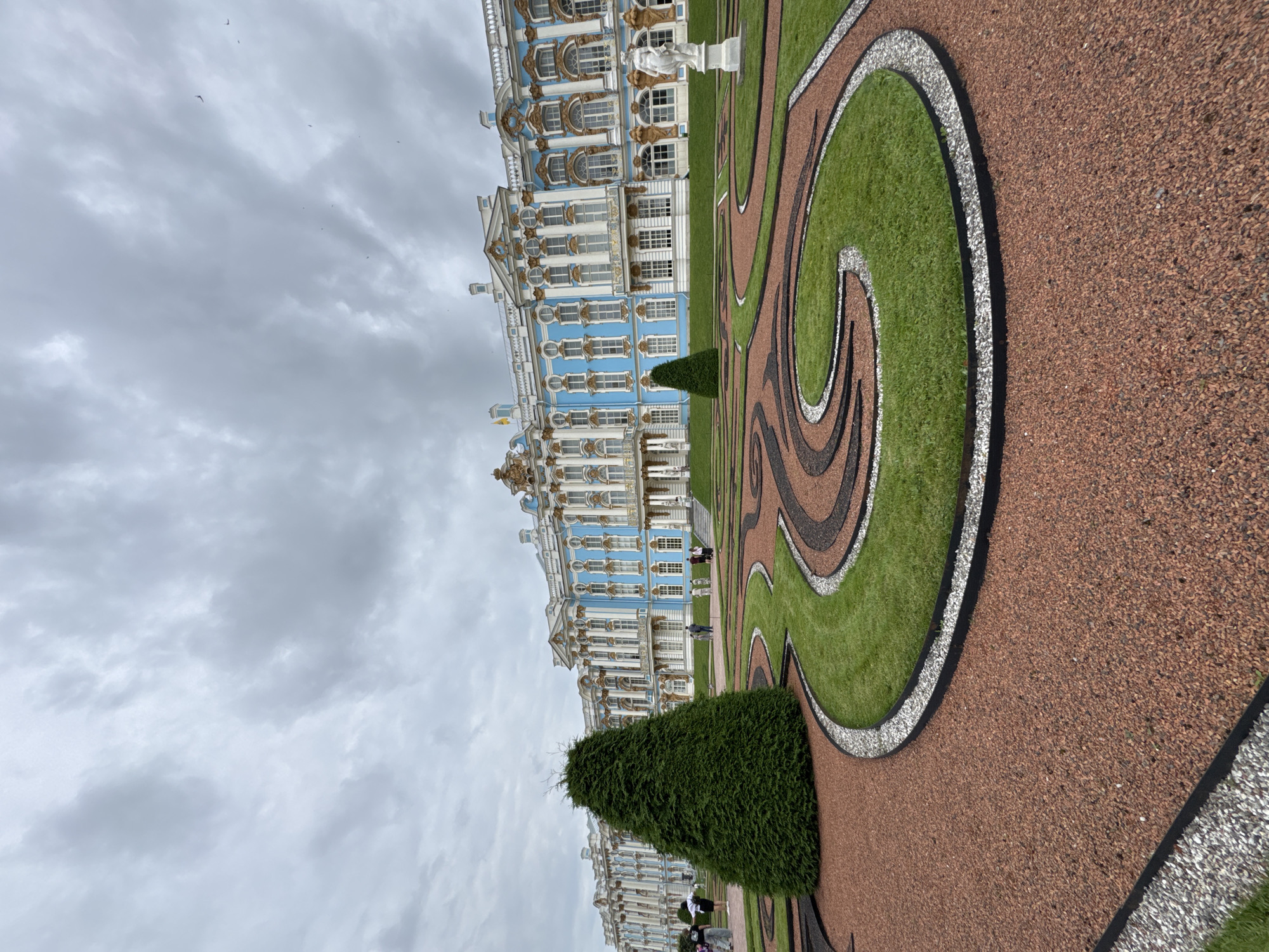 The image shows a wide-angle view of the Catherine Palace in Pushkin, Russia, under a cloudy sky. The palace is a large, ornate building with a distinctive blue facade, white columns, and gold accents. In the foreground, a meticulously manicured garden features swirling patterns of green grass, dark gravel, and reddish-brown gravel, separated by white and black borders. A tall, conical evergreen tree stands to the left of the frame, and a white statue of a woman is visible on the right. Several people are scattered throughout the garden and near the palace entrance, appearing to be tourists. The overall scene depicts a grand architectural landmark and its surrounding formal gardens.