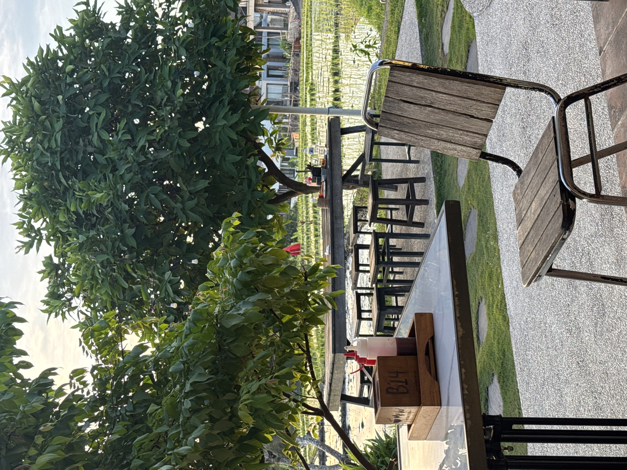 The scene captures an outdoor dining area in North Kuta, Indonesia. A wooden chair with a metal frame is partially visible in the foreground. A glass-topped table is adjacent to the chair, holding a condiment holder labeled "B14" containing sauce bottles. Beyond the table are more tables and chairs, aligned along a path. A lush tree is on the left side of the frame, partially obscuring the view. In the background is a rice field, and some buildings are visible further out. The ground is paved with irregular stone slabs interspersed with patches of grass. The lighting suggests it is daytime.