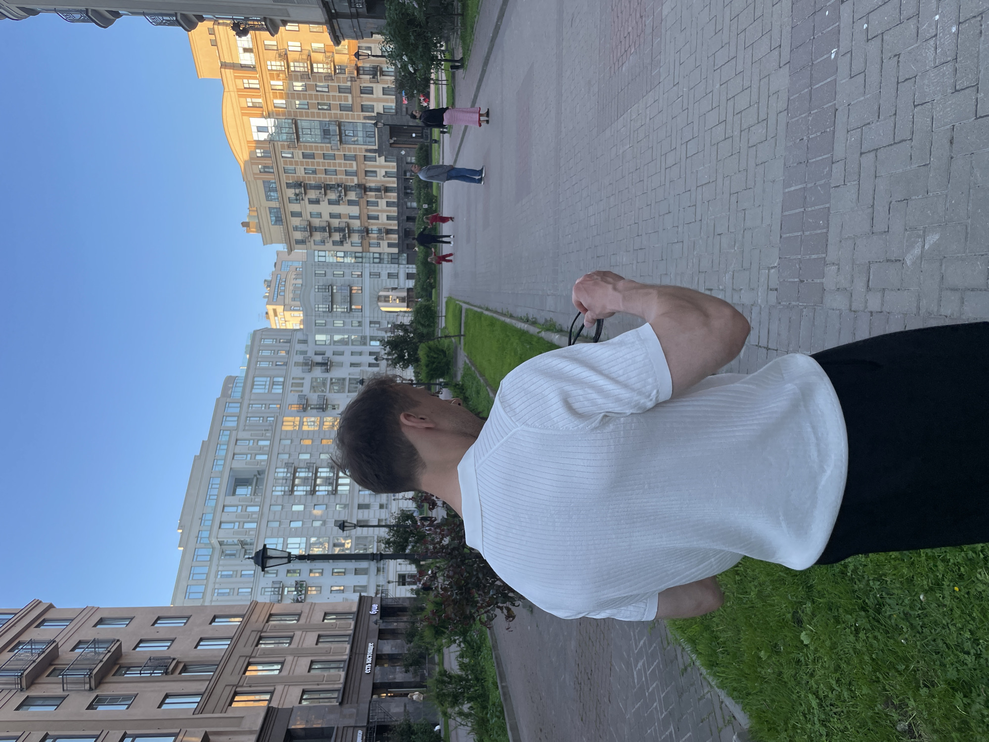 The image depicts a paved pedestrian walkway flanked by green grass and modern multi-story residential buildings under a clear blue sky, likely in Saint Petersburg, Russia.
In the immediate foreground, a man is observed from behind, facing away from the viewer and slightly to the right. He has short brown hair and is wearing a white, short-sleeved polo shirt with vertical ribbing and black pants. His right arm is bent, and he is holding a pair of dark glasses, looking downwards at them.
Further down the path, several individuals are visible. On the left side of the path, two small children, both dressed in red, are standing or playing, with an adult male nearby, wearing a dark top and jeans. On the right side of the path, a woman in a dark top and a long pink and white striped skirt is standing.
The walkway features a herringbone brick pattern with sections of grey and reddish-brown paving stones. Green grassy areas with small bushes and trees line both sides of the path. Several black ornate street lamps are spaced along the walkway.
The background is dominated by various residential buildings. On the left, a light brown multi-story building with balconies is visible. In the center and to the right, a mix of modern multi-story buildings with light-colored facades, large windows, and balconies are present. Some windows reflect the warm glow of the sun, suggesting late afternoon or early evening. One building on the far left displays signage with Russian text, including "ЕСТЬ НАСТОЯЩЕЕ" (YEST' NASTOYASHCHEE - "THERE IS REAL") and "БУШЕ" (BUSHE), indicating the location as Russia, specifically Saint Petersburg due to the prevalence of the "Буше" bakery chain.