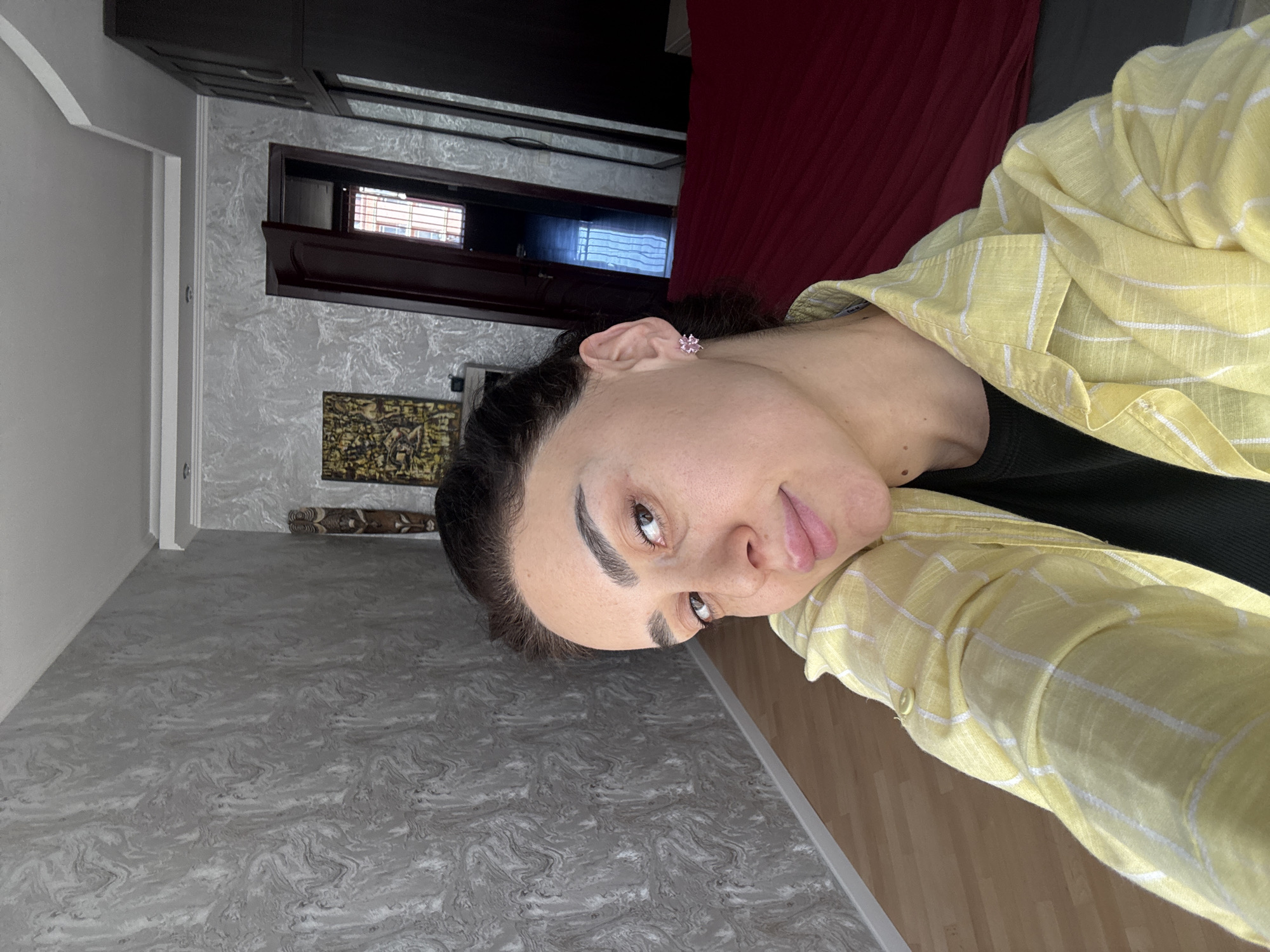 The image shows a woman taking a close-up selfie indoors, in what appears to be a room in Tbilisi, Georgia. She is seen from the chest up, looking directly at the camera with a neutral to slightly content expression. She has dark hair, dark defined eyebrows, and brown eyes. She is wearing a light yellow plaid button-up shirt over a dark top, and a small, light pink flower-shaped stud earring is visible in her left ear.
The background reveals details of the room. Above her head, there's a dark wooden cabinet or shelf. Below it, a textured light grey wall with a wavy or marbled pattern is visible. This wall features a dark wooden window frame with blinds. Further down the wall, a rectangular piece of dark, possibly woven or framed, art hangs. To the left, a white door frame can be seen. The floor in the immediate foreground is a light grey with a similar marbled pattern, transitioning to a lighter wooden floor further back. A rolled-up rug is also partially visible on the floor near the wall art. The room is well-lit, suggesting it is daytime. There is no visible text in the image.