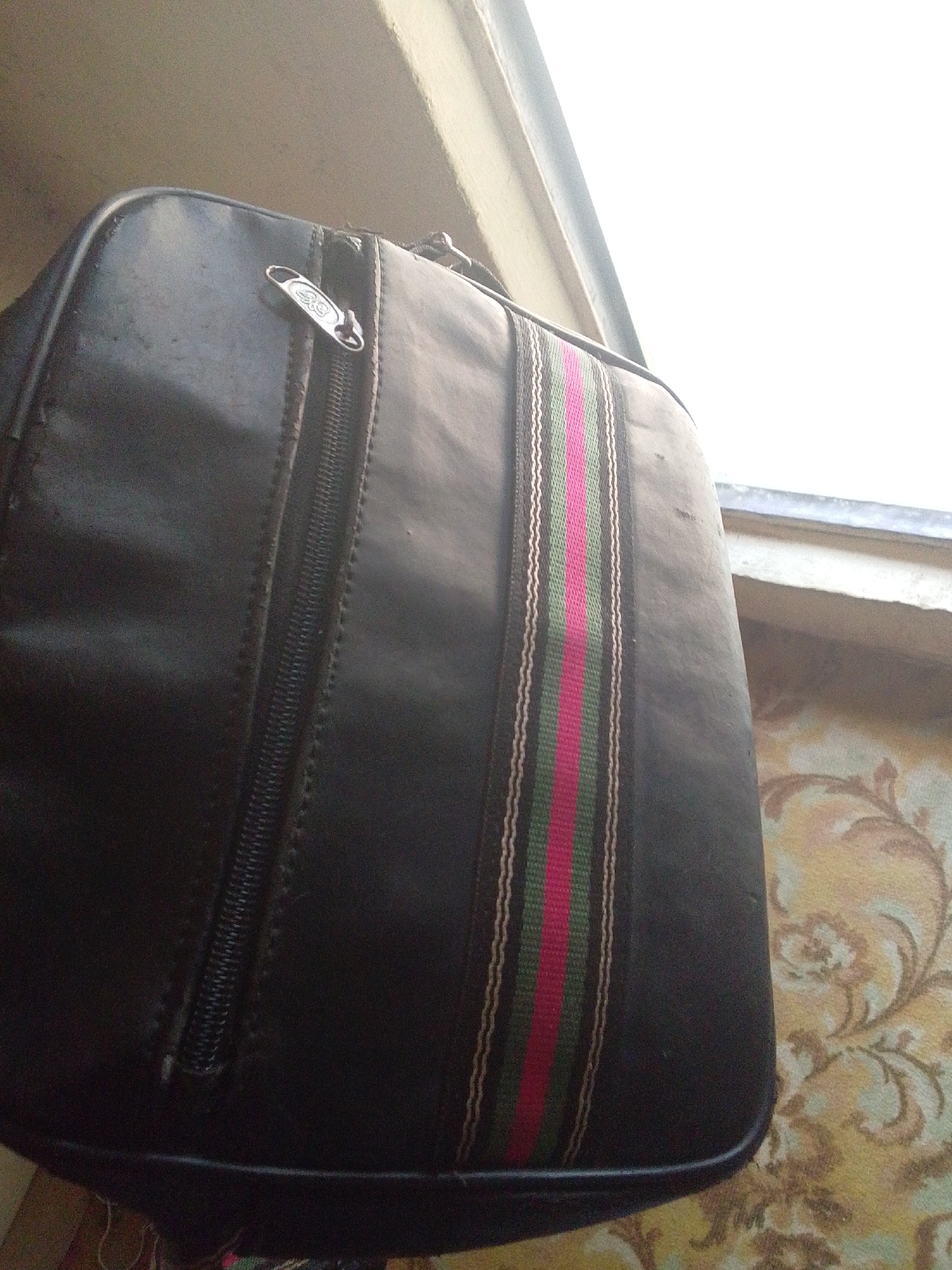 This is a medium-shot, eye-level, close-up photograph of a black duffel bag, positioned slightly off-center to the right. The bag is made of a textured material that appears to be faux leather. A prominent feature is a vertical stripe of fabric running down the right side of the bag, composed of alternating thin white, green, and pink lines. A metallic zipper pull with a circular loop is visible on the upper left portion of the bag. The background consists of an outdoor setting with an overcast sky, suggesting it might be daytime. To the right of the bag, there's a glimpse of a wall covered in patterned wallpaper. The overall lighting is somewhat dim, with the bright sky in the background creating a slight overexposure. No people are visible in the image. The focus is solely on the bag and its immediate surroundings, offering a detailed view of its design and texture. The location is Jalingo, Nigeria.