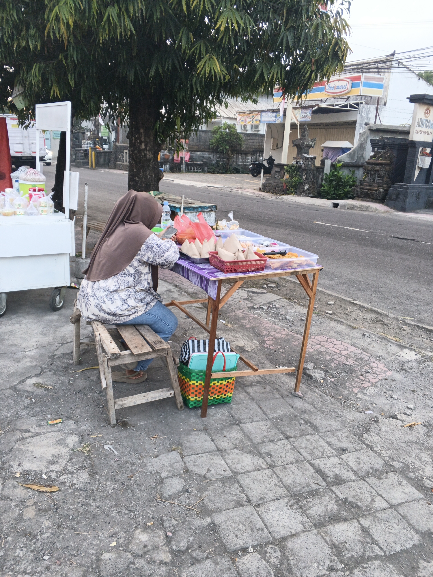 In Sukawati, Indonesia, a woman wearing a brown hijab and a patterned shirt is seated on a wooden stool beside a street food stall. She is holding a mobile phone and appears to be interacting with it. The stall features a table laden with food items, including packaged meals and drinks, some of which are in red plastic bags and clear plastic containers. A red basket containing more food items is also visible on the table. Beneath the table, a colorful woven basket with green, orange, and blue sections holds striped bags. The stall is situated on a paved sidewalk next to a road with a sidewalk on the opposite side. Behind the stall, a large tree provides shade. In the background, buildings, including an Indomaret convenience store, and parked motorcycles are visible. The overall scene depicts a typical street-side food vending operation in an urban setting.