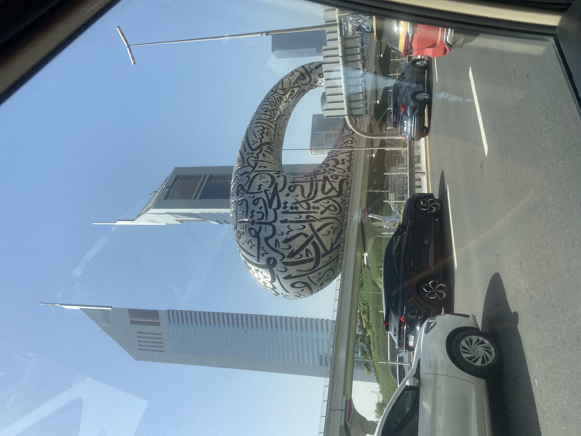 The image, taken from inside a vehicle on a roadway in Dubai, United Arab Emirates, features the Museum of the Future, a large, elliptical building adorned with Arabic calligraphy. Several other buildings and skyscrapers are visible in the background. A section of traffic is seen to the right of the frame. The sky is clear and blue.
