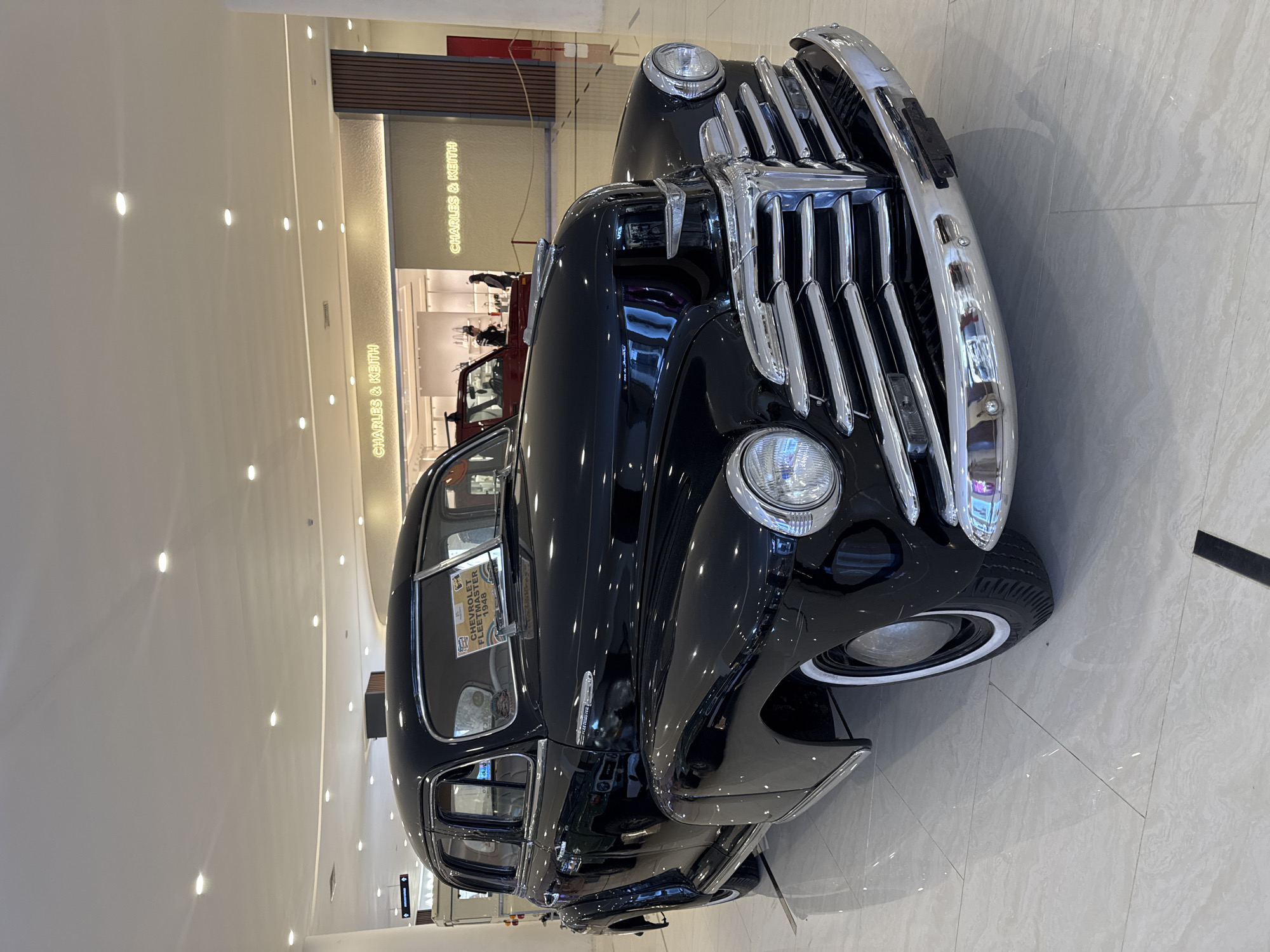 A black 1948 Chevrolet Fleetmaster is on display in the Beachwalk Shopping Center in Kuta, Indonesia. The vehicle's chrome grille and headlights are prominent in the foreground. In the background is a storefront for "CHARLES & KEITH." The floor is polished tile.
