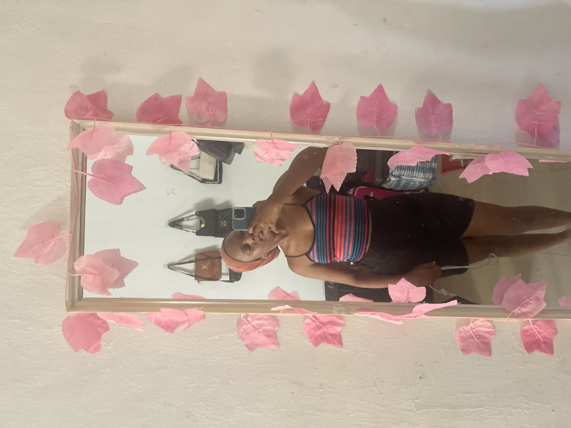 In a room in Jos, Nigeria, a young woman with a light brown headscarf and a striped tank top poses for a selfie in a full-length mirror. The mirror is framed with a string of artificial pink leaves. Behind her, several handbags are hanging on the wall. The room has white walls and a wooden floor.