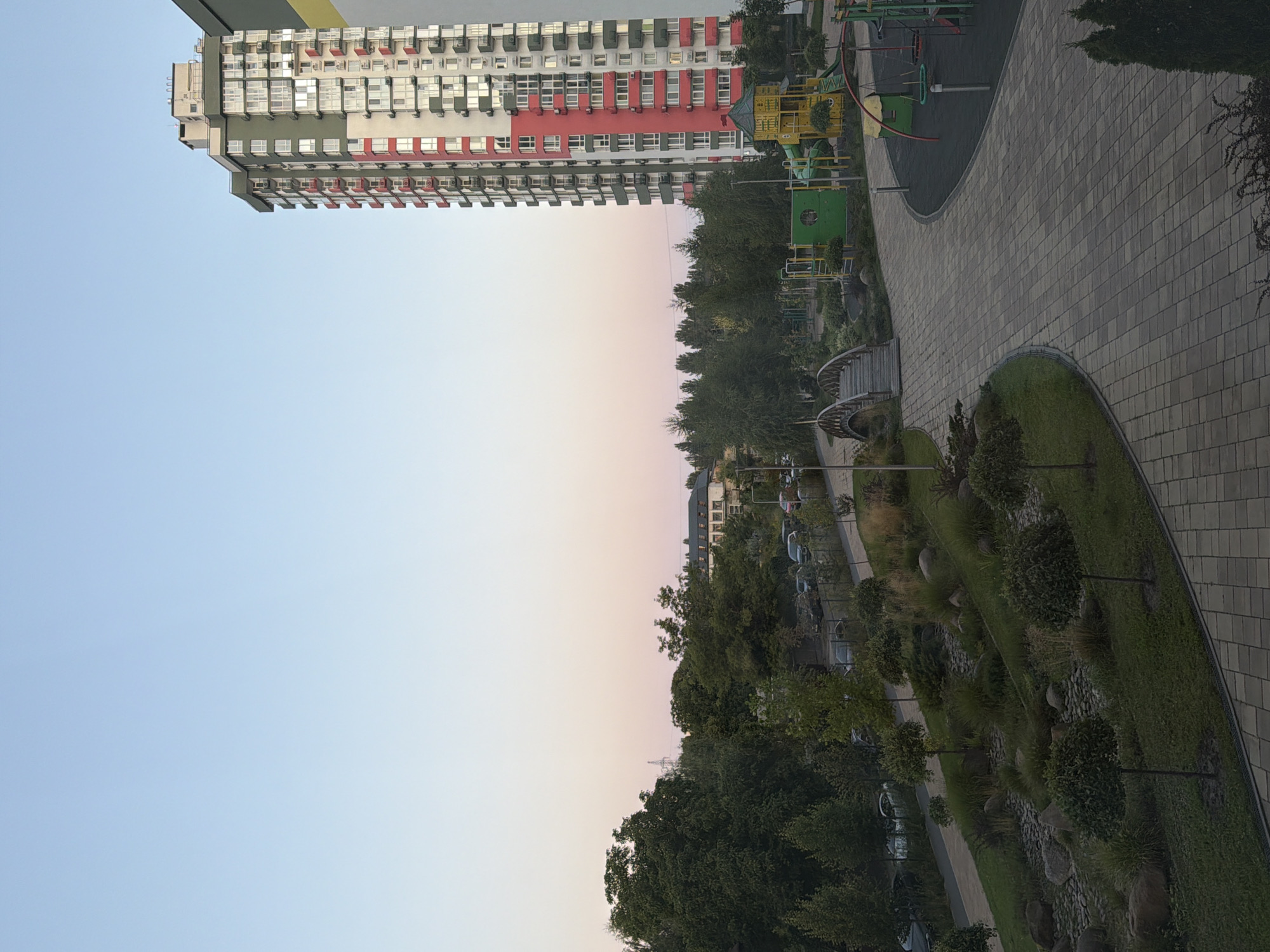 The image shows a modern residential building in Kyiv, Ukraine, with a playground and landscaped areas in the foreground. The building is tall and multi-story, with a facade featuring a combination of white, red, and green panels. The playground includes various structures like slides, swings, and a climbing frame, all set on a rubberized surface. A paved walkway curves through the scene, connecting to the playground and a small, arched wooden bridge that spans a decorative, rocky stream bed with manicured trees and shrubs. In the background, other buildings and a line of trees are visible, along with parked cars along a street. The sky is clear and light blue with a hint of pink near the horizon, suggesting it is either early morning or late evening.