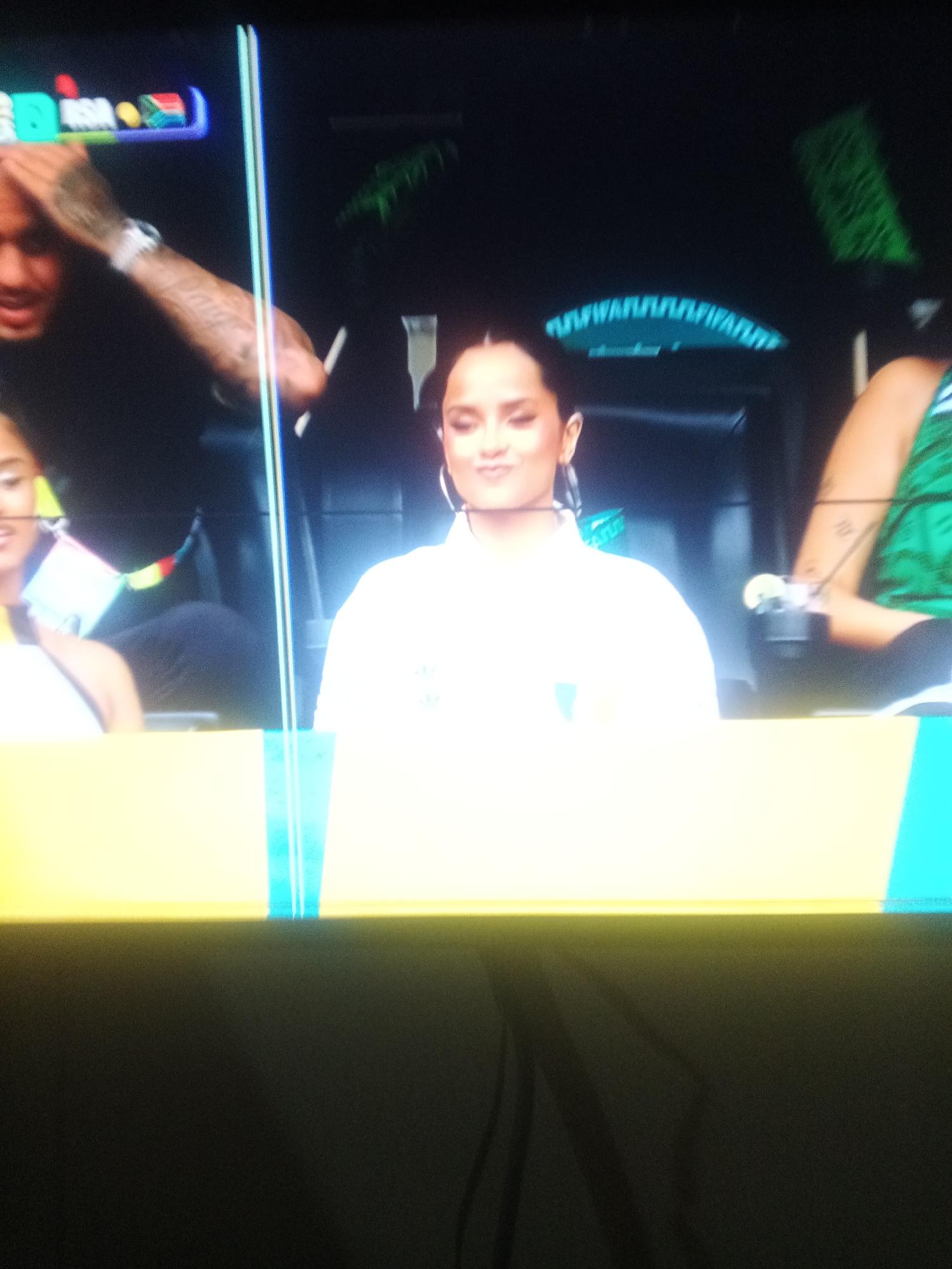The image captures a scene that appears to be a broadcast or live event, likely from Jalingo, Nigeria, given the prompt. In the foreground, a person with dark hair, wearing a white shirt with a subtle logo, is seated and looking slightly downwards with a relaxed expression. Their face is illuminated, suggesting they are under stage or studio lighting. To their left, another person, partially visible, is also seated. To the right of the central figure, another individual's arm is visible, showing tattoos and a watch. In the background, there are elements that suggest a set or stage, including what looks like a neon sign with a wave-like pattern and some greenery. The overall lighting is artificial, and the image quality suggests it was taken with a camera at a distance or under challenging light conditions. The yellow and cyan bars at the bottom indicate a separation or graphic overlay, common in broadcast media.
