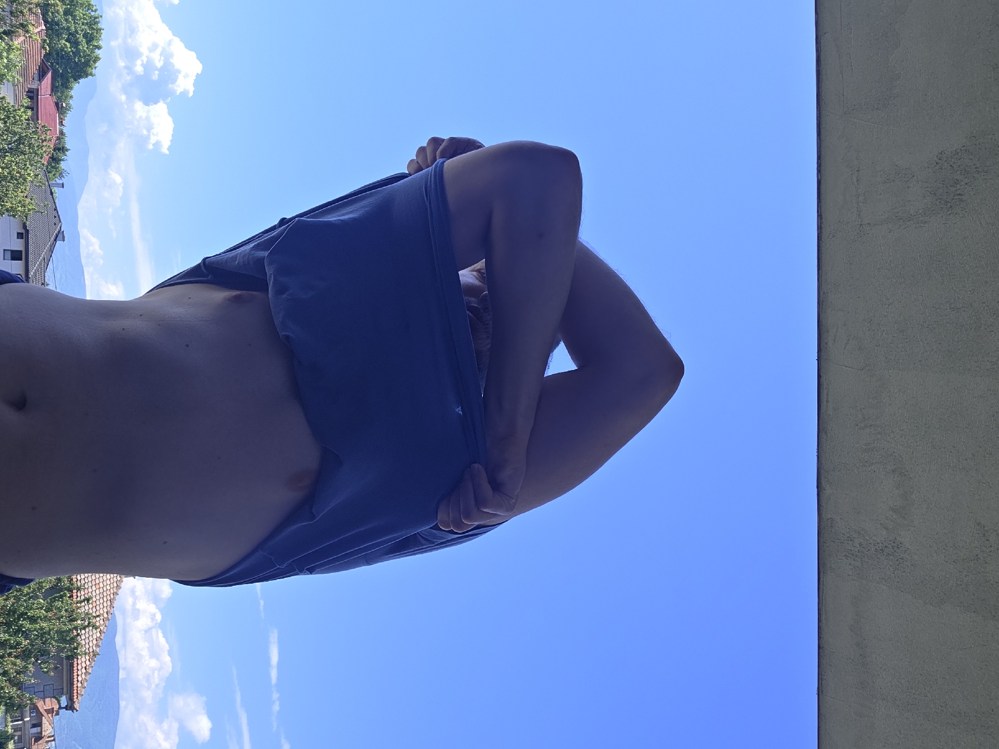 A person is lifting a blue T-shirt up, revealing their abdomen. The setting appears to be outdoors on a sunny day, evidenced by the clear blue sky. In the background, trees, houses with red roofs, and mountains are visible, suggesting a location in Yagodovo, Bulgaria. A concrete wall is on the right side of the frame.