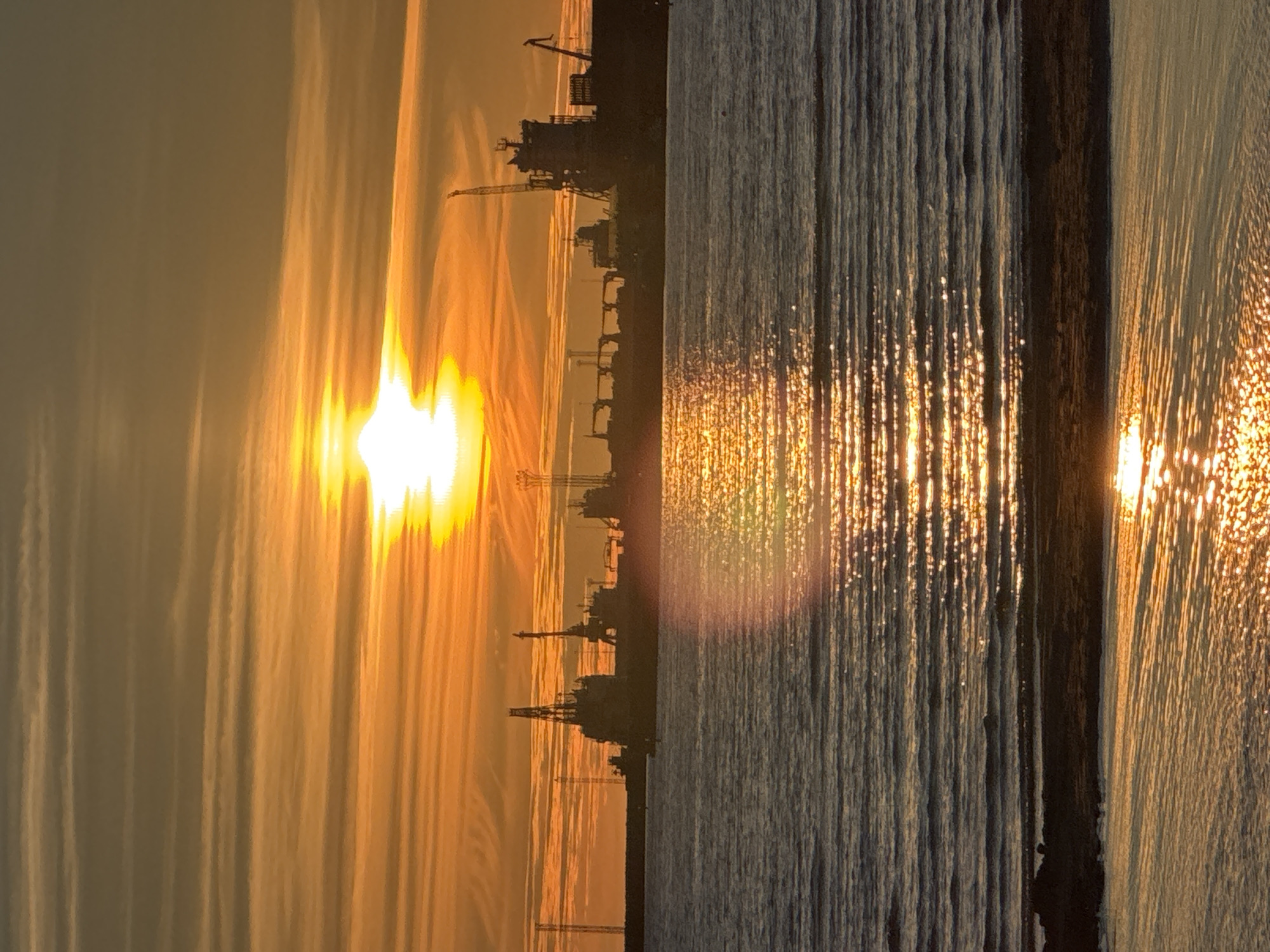 The image depicts a harbor scene at sunset in Tallinn, Estonia. The sun, a bright orb, is setting on the horizon, casting a warm orange and yellow glow across the sky and the water. Wispy clouds stretch across the sky, catching the sunlight. In the background, silhouetted against the vibrant sky, are several large ships and industrial cranes, suggesting a working port. The water in the foreground reflects the sunset, creating shimmering patterns. A small stretch of dark, wet sand or shoreline is visible at the bottom of the image. The overall impression is one of a serene and industrial landscape bathed in the dramatic colors of a sunset.