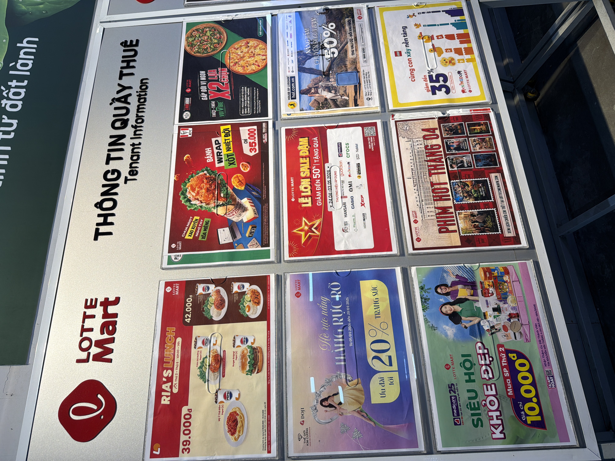 The image shows a display case with various advertisements for Lotte Mart, a South Korean retail chain, likely located in Đà Nẵng, Vietnam. The ads promote sales and promotions from various brands and departments within Lotte Mart, including: Ria's Lunch deals, pizza, KFC wraps, Doji jewelry, house of luggage, skincare, a movie promotion, and Lego toys. The ads are colorful and visually appealing, targeting local Vietnamese consumers.