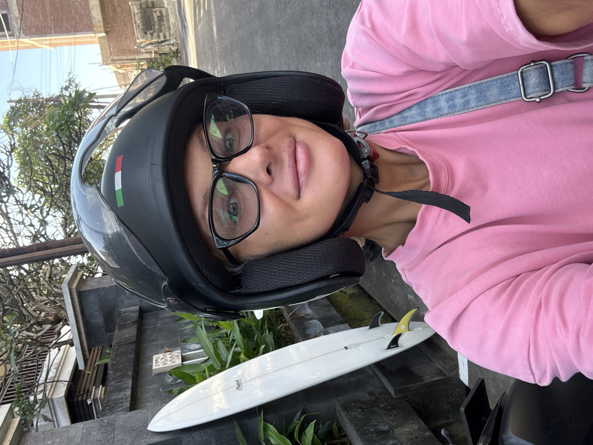 A woman wearing a black helmet with an Italian flag sticker and glasses is looking at the camera. She is wearing a pink long-sleeve shirt and a blue denim strap. To her side, a white surfboard with black and yellow fins is visible. The background includes a street scene with buildings and greenery. The location appears to be a city, with a combination of residential or commercial buildings and tropical vegetation. The specific city and region are unknown.