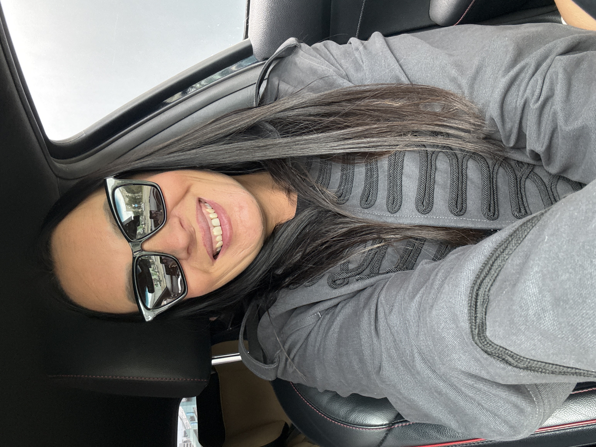 A woman wearing sunglasses and a grey jacket with ornate dark trim is sitting in the driver's seat of a car. She has long, dark hair and is smiling. The interior of the car is visible, with black leather seats and red stitching. The background is blurred, but suggests an outdoor environment. It is not possible to determine the location in Izhevsk, Russia from the image.