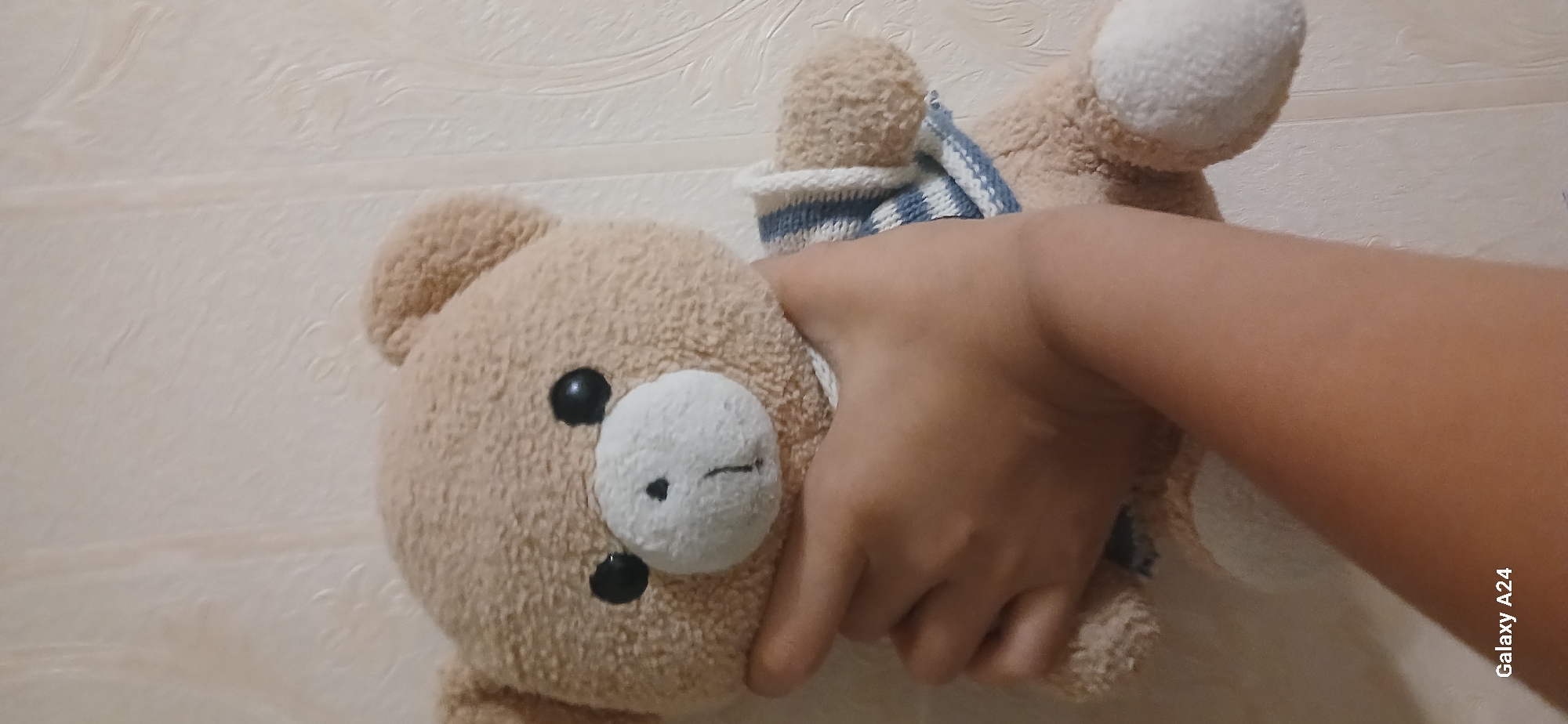 A hand holds a light brown teddy bear against a textured, off-white wall. The teddy bear has black eyes, a white muzzle with a black nose and a stitched mouth. It is wearing a striped sweater with blue and white stripes. The hand is positioned on the bear's chest and lower face. The overall lighting is dim. There are no discernible elements that suggest the location is in Bishkek City, Kyrgyzstan.