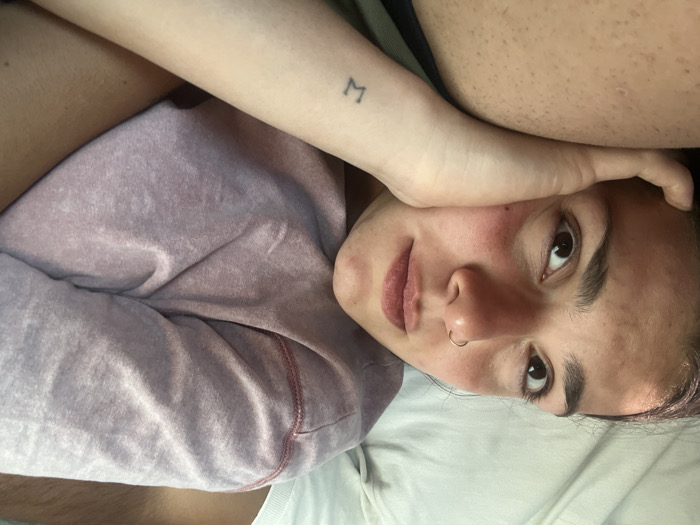 A person lies sideways on white bedding, gazing upwards with a calm expression. Their left arm rests on their forehead, revealing a small tattoo of the letter 'M' on the wrist. They have brown eyes, a nose ring, and flushed cheeks. A light purple sweatshirt is visible on their left side. The background shows blurred skin and clothing, possibly indicating someone lying beside them.