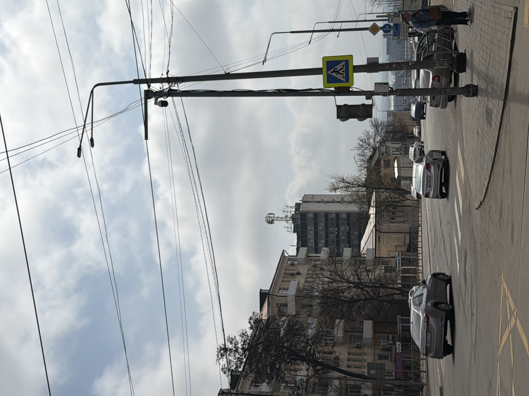The image depicts a street scene in Rostov-on-Don, Russia. The street is lined with buildings of varying heights and architectural styles, including an older building with multiple windows and a taller, more modern structure with visible antenna structures on its roof. Overhead, there are numerous power lines and a pedestrian crosswalk sign. Cars are parked along the street and driving along the street. A pedestrian is standing on the sidewalk near the crosswalk, wearing a jacket and carrying a bag. The sky is overcast, suggesting a cloudy day. The location may be near the intersection of Bolshaya Sadovaya Street and Budyonnovsky Prospekt, based on the architecture and street signs.