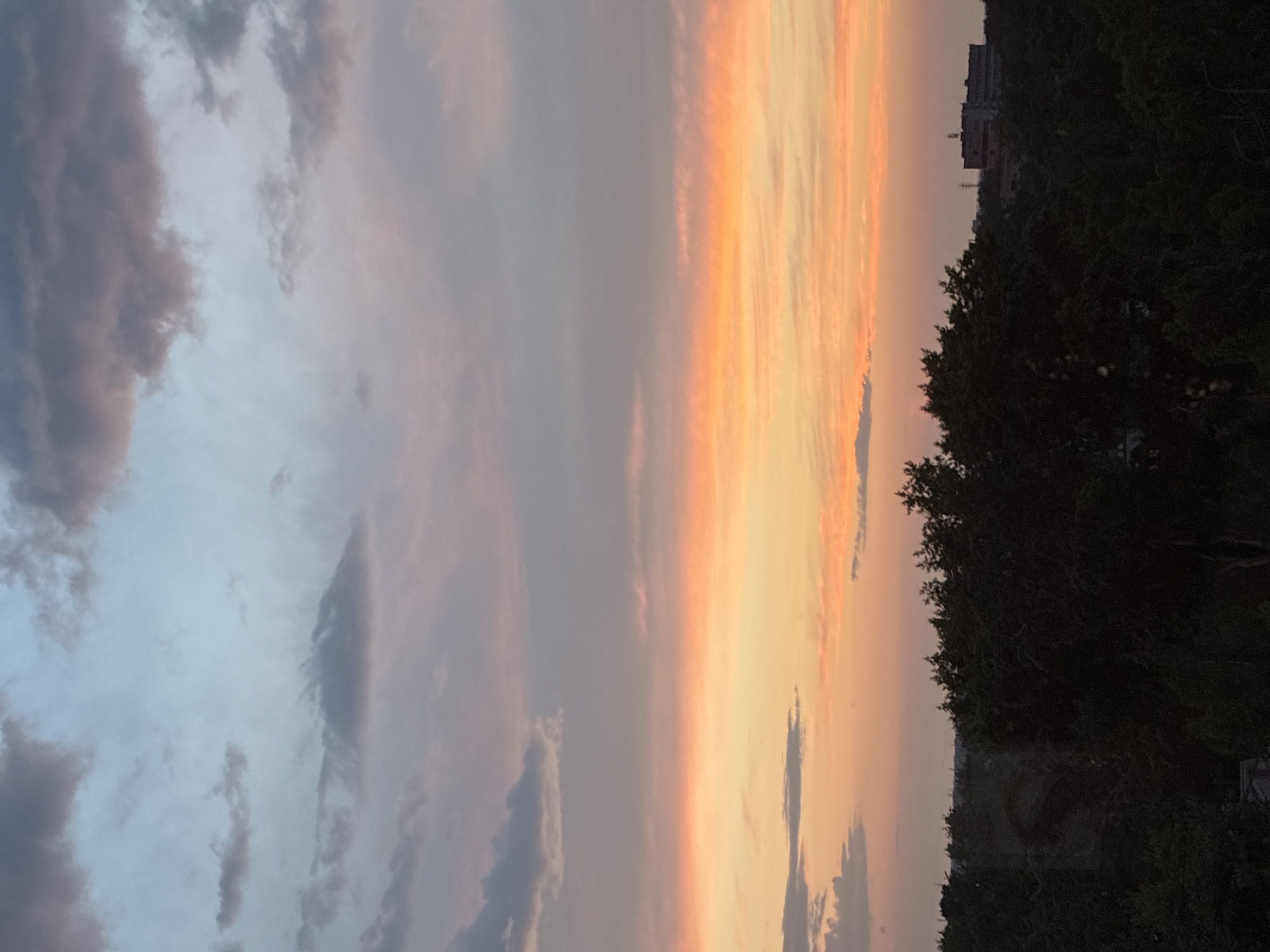 The image depicts a dusky sky with vibrant hues of orange and pink, transitioning to a pale blue at the top, indicating either sunrise or sunset. Fluffy clouds, tinged with grey and purple, are scattered across the sky. Below the sky, a dense line of dark green trees forms a silhouette against the colorful horizon. In the distance, beyond the trees, a building with multiple windows and a tower-like structure is visible. The overall scene suggests an outdoor setting in a semi-urban or suburban area at dusk or dawn. The location is identified as Unknown, Unknown.