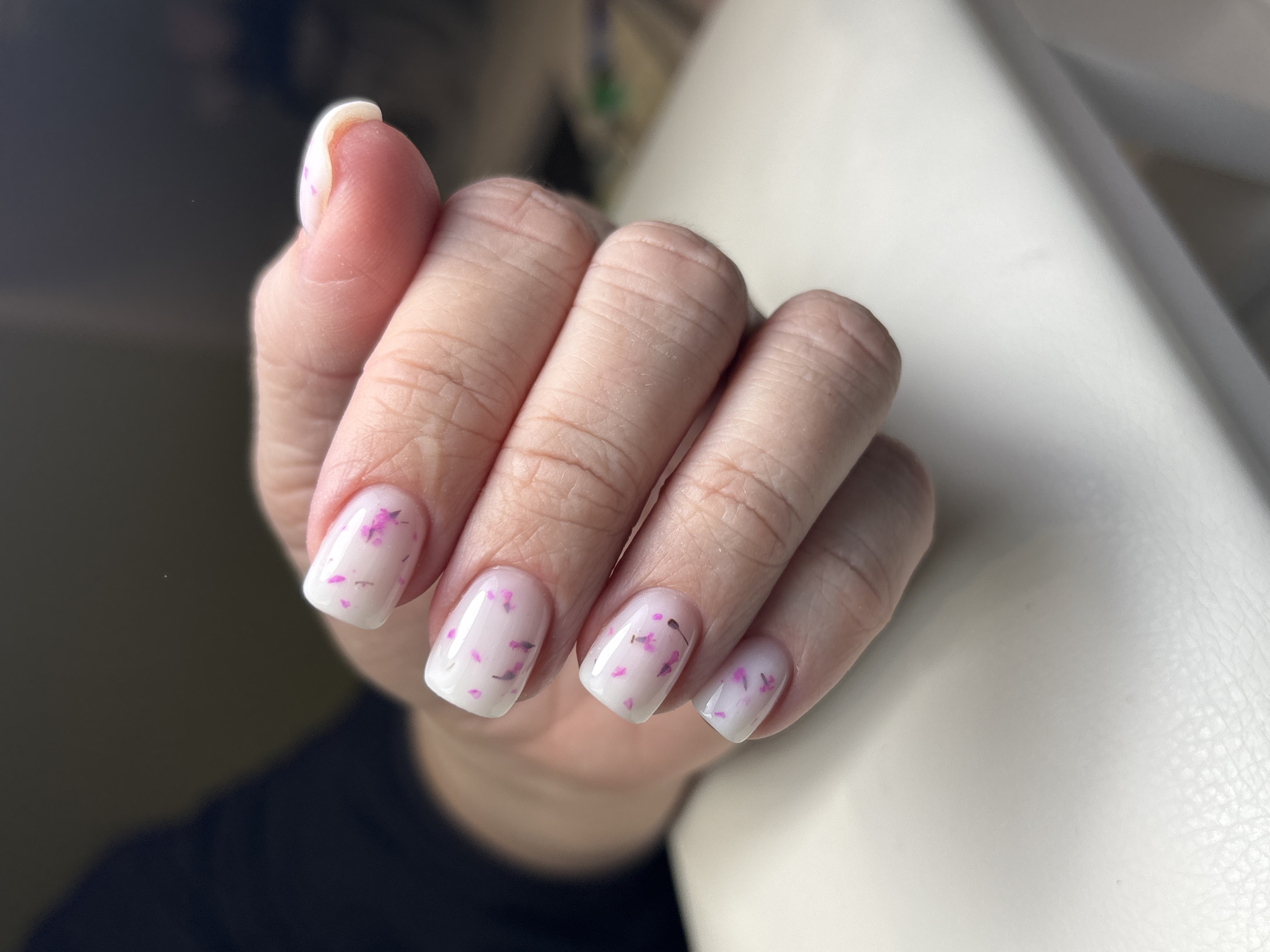 A close-up captures a person's left hand with manicured nails. The nails are painted with a light, milky color and decorated with small, pink and black floral pieces. The hand is positioned with fingers slightly curled, resting against a light-colored, textured surface. In the background, a dark color provides contrast, suggesting an indoor setting. No location is specified.