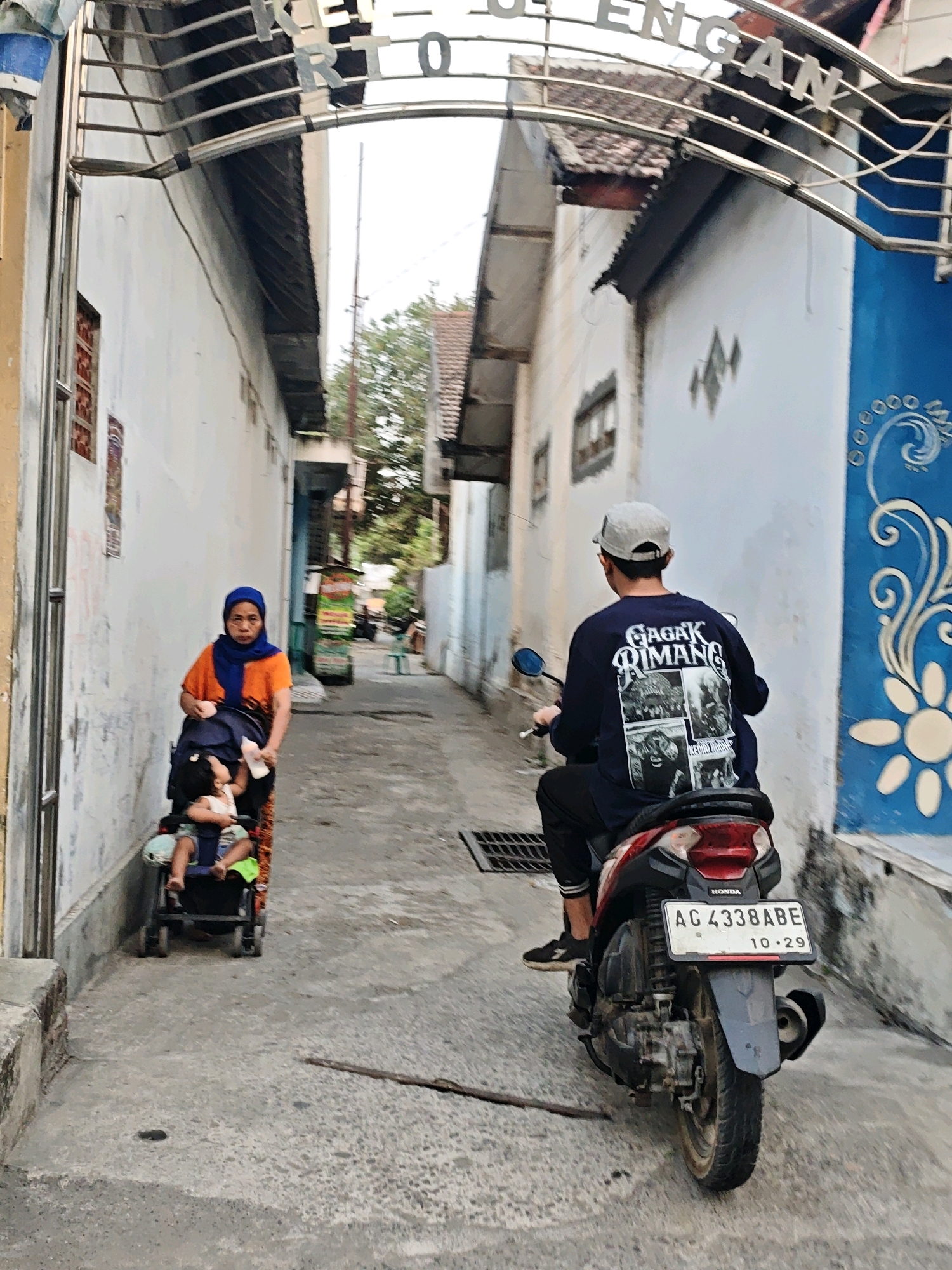 In a narrow alleyway in Kediri City, Indonesia, a person on a red and black motorcycle rides away from the camera. The rider wears a dark long-sleeved shirt with graphics on the back and a light-colored baseball cap. The motorcycle has a license plate that reads "AG 4338 ABE 10.29". To the left, a woman wearing an orange shirt and a blue hijab pushes a stroller containing a baby. The alley is lined with white buildings, one of which has a vibrant blue facade with a white floral design. The ground is paved with concrete. Above the alley, a metal archway with lettering is visible.