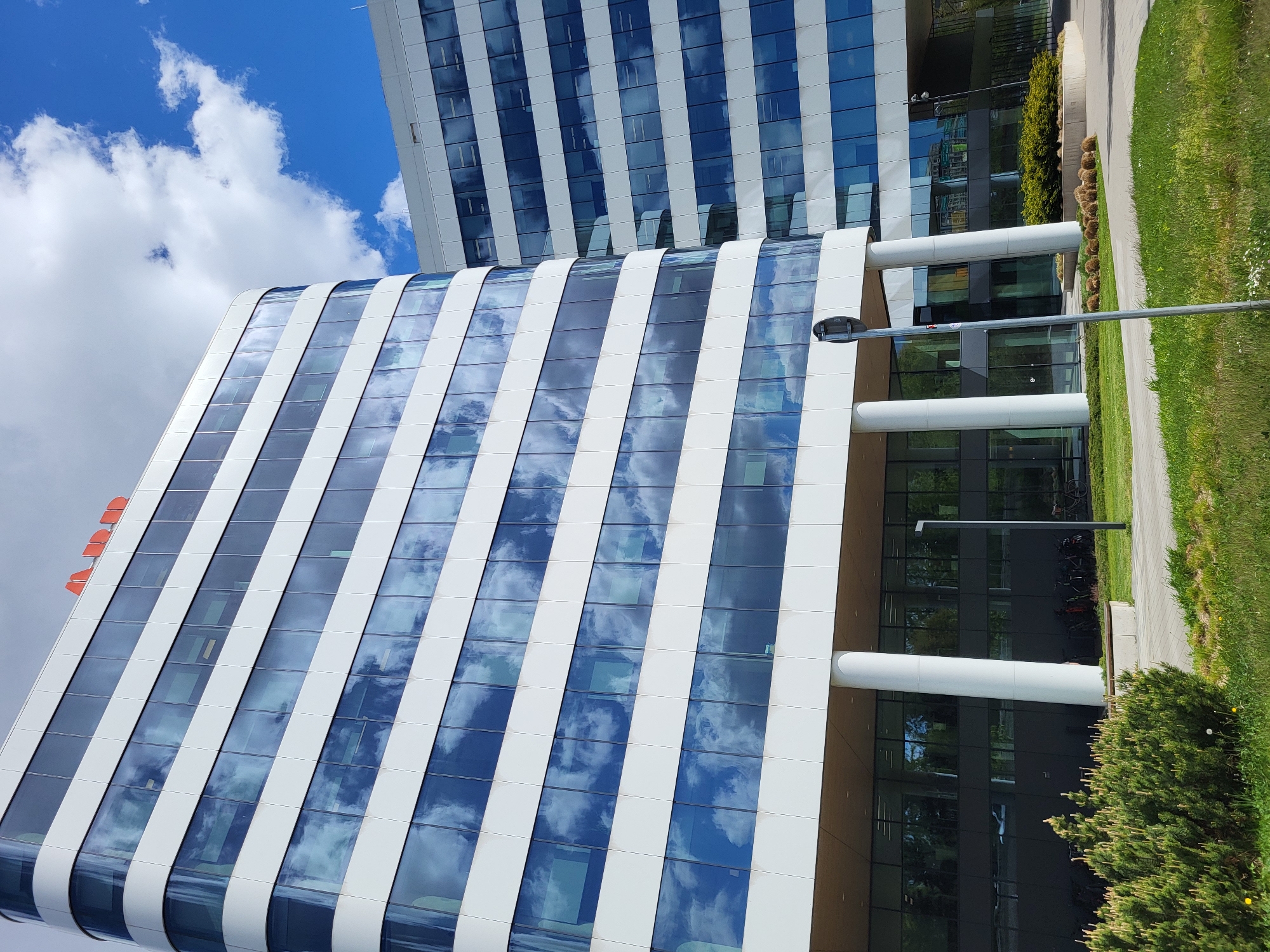 A modern architectural scene, captured on a bright, sunny day, features two contemporary buildings under a clear blue sky adorned with white clouds. The prominent building in the foreground, on the left, boasts a curved facade composed of alternating vertical white structural bands and highly reflective, blue-tinted glass windows. These windows vividly mirror the sky and clouds, integrating the structure with its environment. Small red vent-like features are visible on its roof. Behind it, a second building with a similar modern aesthetic, but with a straighter, more traditional rectangular form, also reflects the expansive sky.
At the ground level of the foreground building, large white cylindrical pillars are visible. The lower sections of the buildings feature dark, reflective windows, subtly revealing interior spaces and a bicycle rack with parked bikes, suggesting an active workplace. The surrounding area includes a paved sidewalk and well-maintained green grass, interspersed with neatly trimmed bushes and smaller shrubs. A tall, slender street light stands towards the middle-right of the scene. The weather is bright and clear, indicating a pleasant daytime setting. No people are visible, and there is no discernible text. The image conveys a sense of sleek design and an orderly urban landscape.
