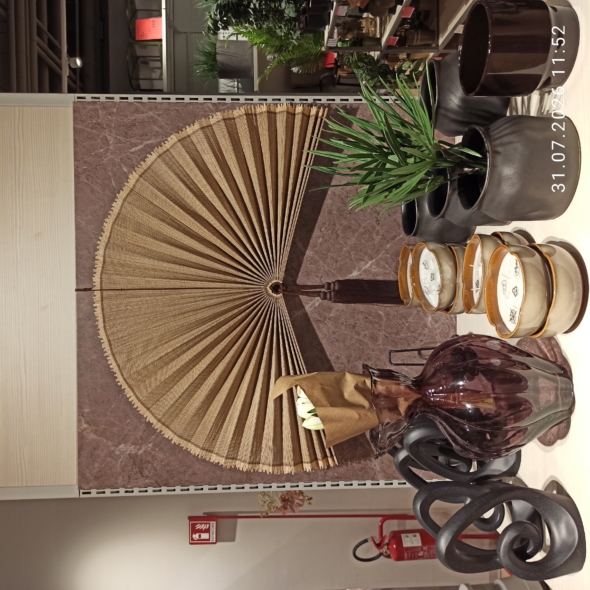 A close-up shot captures a display of decorative items and plants on a white shelf, likely in a retail setting. On the left, a red fire extinguisher is mounted on a white wall. In the center, a large, fan-shaped woven wall hanging with a tassel dominates the background. In front of it, a dark purple glass vase holds a bouquet of white tulips wrapped in brown paper. To the right of the vase, a stack of seven beige ceramic bowls containing white candles is arranged. Further to the right, a potted green plant with long, spiky leaves sits in a dark gray ceramic pot, flanked by two other similar pots and a dark brown ceramic planter. The scene is well-lit, highlighting the textures and colors of the objects. A timestamp in the bottom right corner reads "31.07.2026 11:52". The context of Modugno, Italy, is provided as the location.
