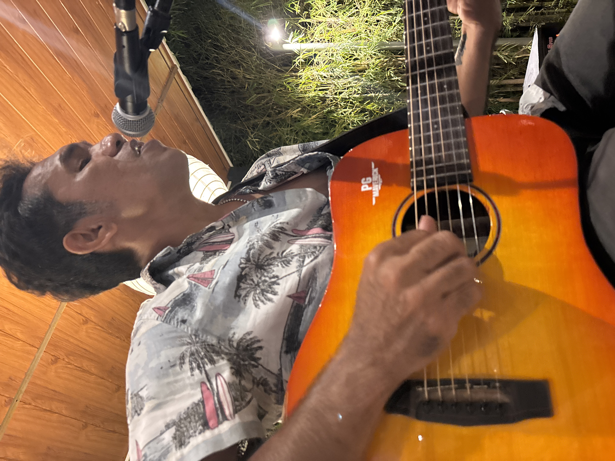 A man plays an acoustic guitar and sings into a microphone in front of a bamboo fence and a light fixture. The man is wearing a light-colored shirt with a pattern of palm trees and surfboards. The guitar is orange and yellow. The scene appears to be outdoors, possibly at night, given the lighting. The location is Kuta Selatan, Indonesia.