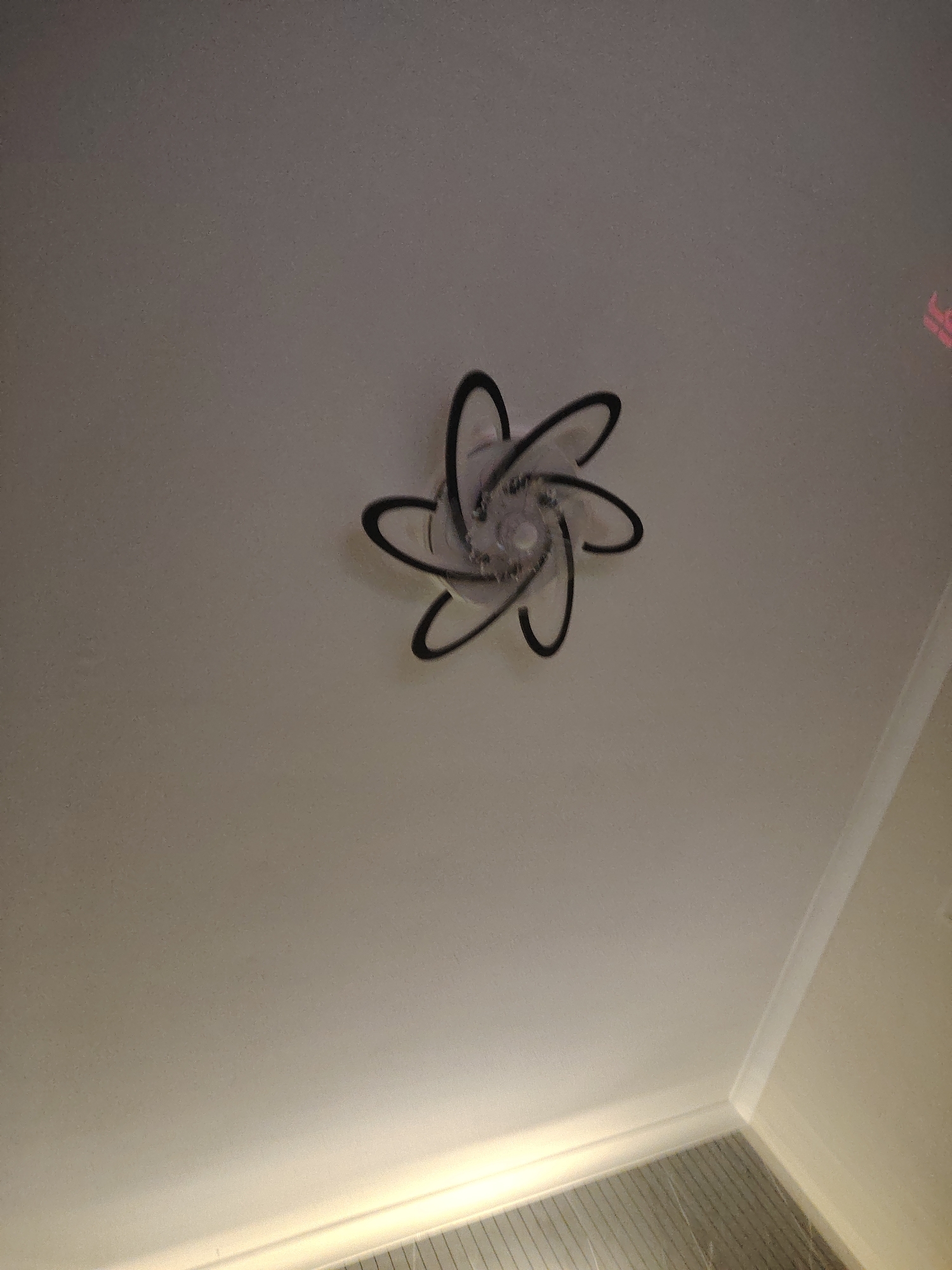 A ceiling light fixture is centrally located on a white ceiling. The fixture has a spiral or starburst design with six curved, black arms. The arms appear to be made of metal. The lighting on the ceiling is dim, casting a faint glow. In the upper right corner of the image, a portion of a pink neon sign is visible, with the letters "LO" partially discernible. The rest of the image shows a plain white ceiling and a corner of the room where the ceiling meets the wall with white molding. The floor is not visible. The image does not provide any specific details about the city of Huntingdonshire, United Kingdom, or any people or actions taking place.