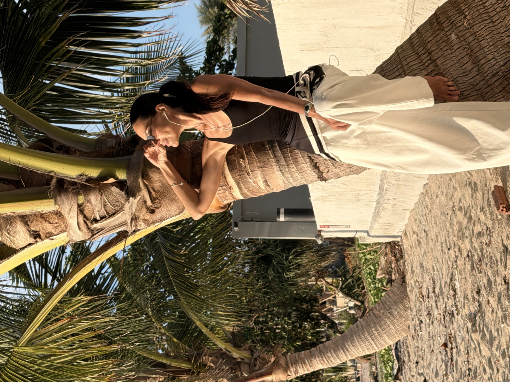 A woman wearing a black tank top, white pants, and sunglasses sits leaning against a palm tree trunk on a sandy beach. She has white earbuds in and is looking to her right, with her hand near her face. The palm tree is large and textured, with green fronds extending above and to the left. In the background, there is a light-colored wall with a dark cylindrical object on it, and beyond that, lush green foliage. The sand is light brown and scattered with small debris. The scene is illuminated by warm, golden light, suggesting either sunrise or sunset. The location is identified as Ko Samui District, Thailand.