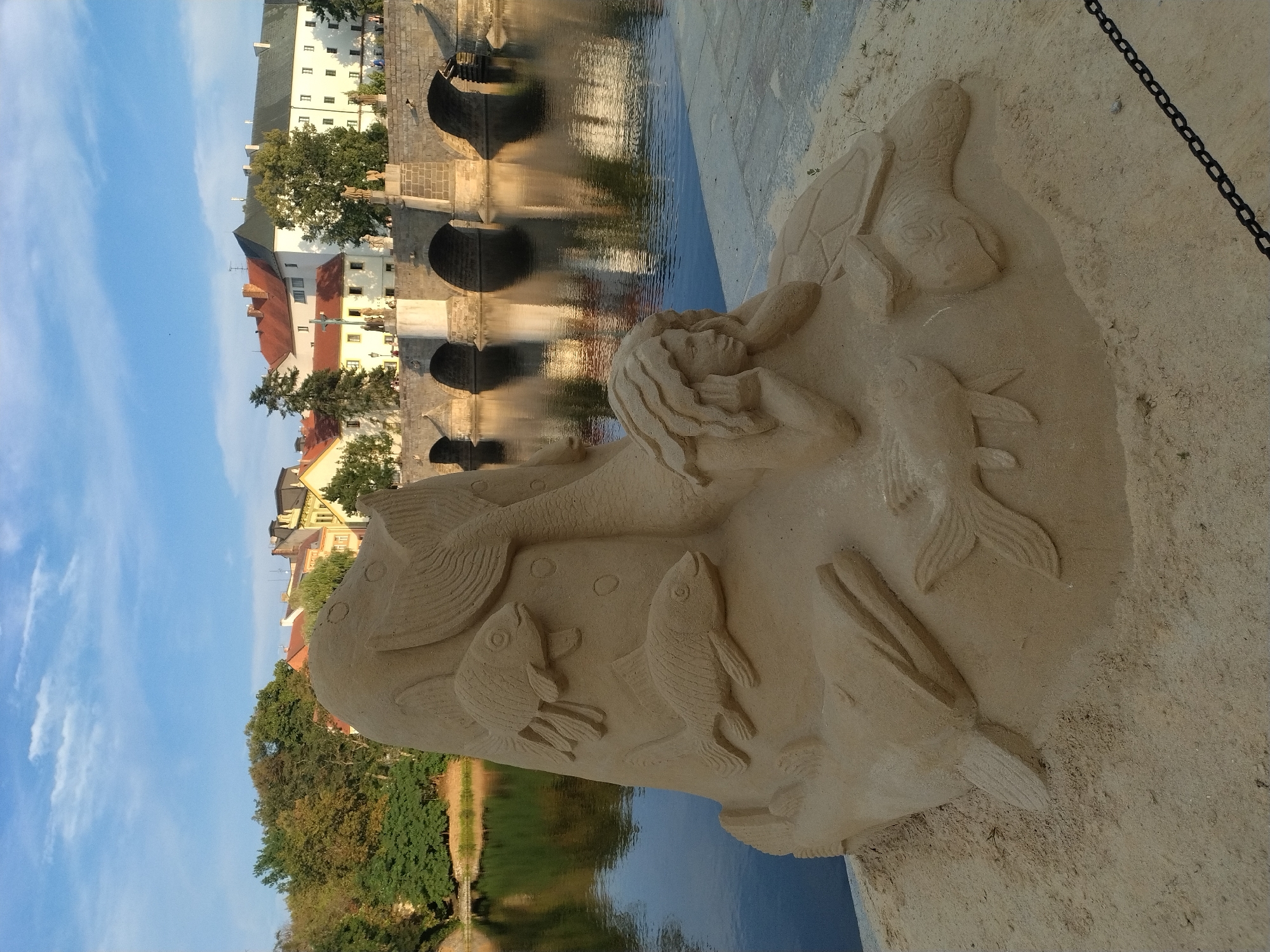 An elaborate sand sculpture of a mermaid and marine life sits on a sandy embankment next to a body of water, likely the Otava River in Písek, Czechia. The sculpture depicts a mermaid with flowing hair resting her head on her hands, surrounded by fish and a sea turtle. Behind the sculpture, a stone bridge with multiple arches spans the water, and beyond that, buildings with red and yellow roofs line the opposite bank under a blue sky with scattered clouds. The water reflects the bridge and the buildings. The foreground shows a textured sandy surface with a black chain border.