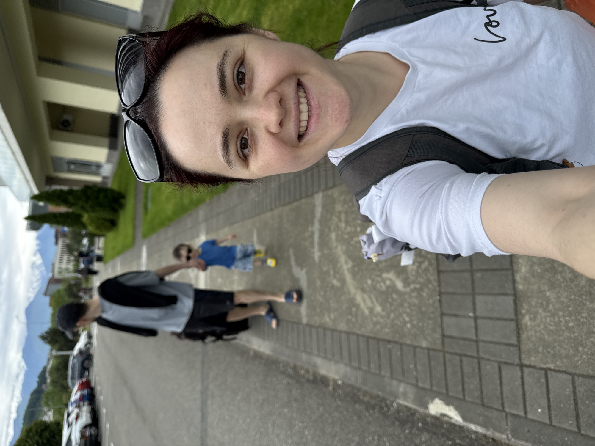 A medium shot of a Caucasian woman smiling and looking towards the camera, which is at the top of the frame, with sunglasses on her head, wearing a white T-shirt with black writing, and a gray backpack. Behind her on a sidewalk of gray pavers and gray concrete is a man with a baseball cap in a gray shirt and black shorts walking behind a child in a blue shirt and blue shorts wearing sunglasses. Behind them are parked cars and in the distance are buildings, trees, and mountains with cloudy skies. The scene is a street scene in Sochi, Russia.