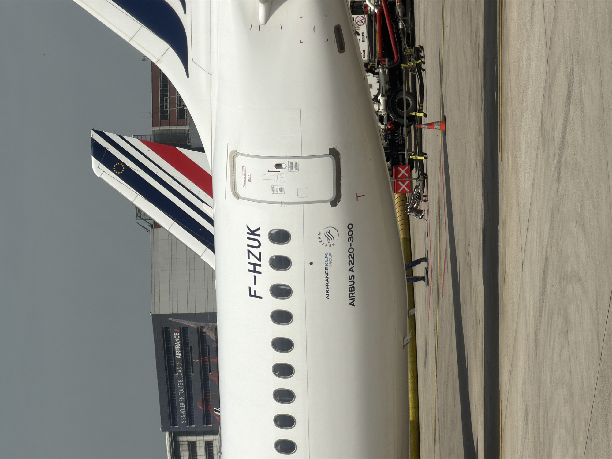 The image shows the rear section of an Air France Airbus A220-300, registration number F-HZUK, parked on the tarmac at a location implied to be in or near Le Mesnil-Amelot, France, given the Air France building and livery on the aircraft. A building displaying the Air France logo with the phrase "S'ENVOLER EN TOUTE ÉLÉGANCE. AIRFRANCE" is visible in the background. The aircraft has an emergency exit door, a tail with blue and white stripes, and another tail with red, white, and blue stripes. Servicing equipment, including what appears to be a refueling or maintenance vehicle, is alongside the aircraft. A person's legs are visible near the servicing equipment.