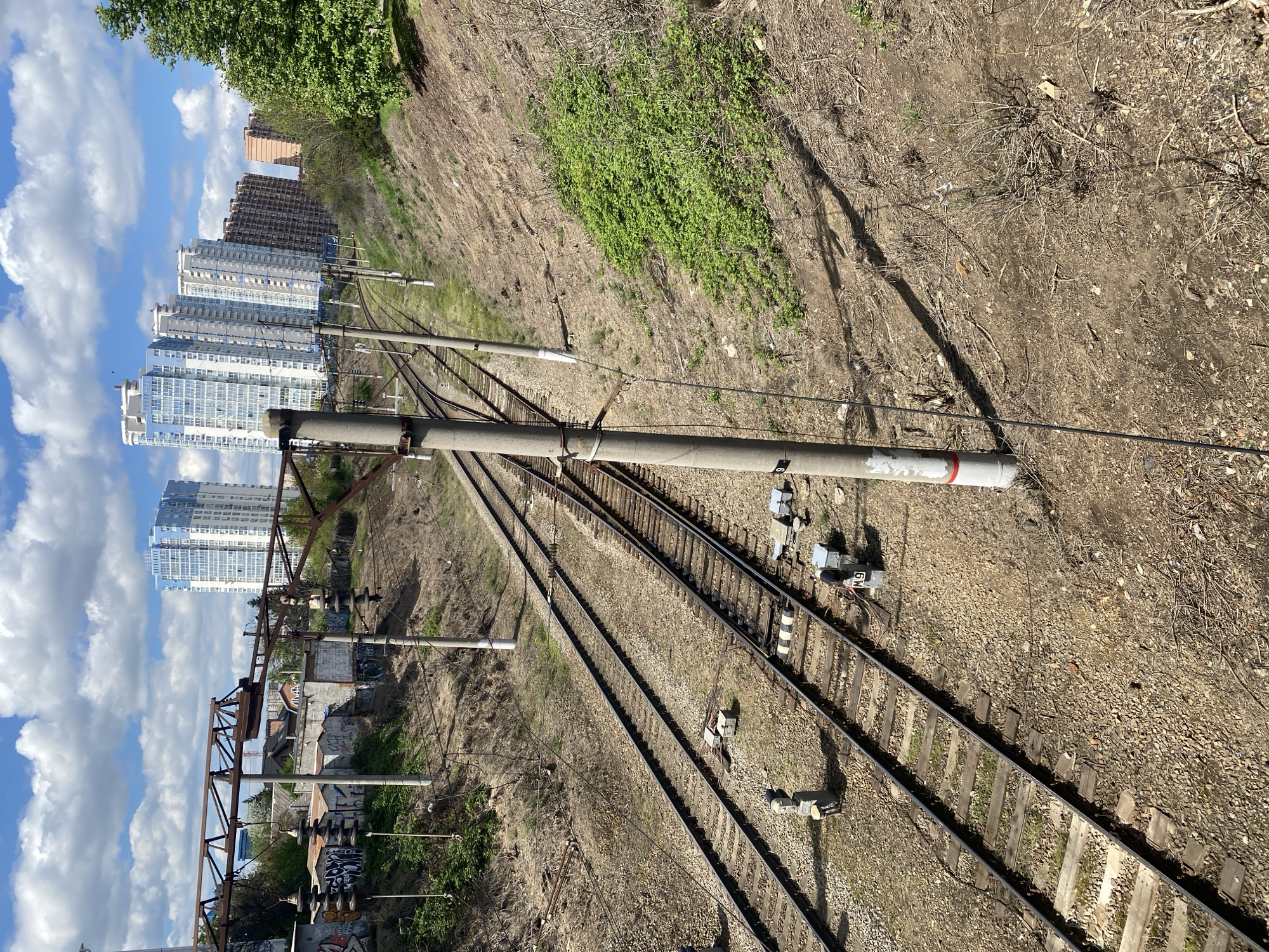 The image shows a railway track in an urban environment. Two sets of tracks run parallel, with gravel ballast visible between and around the rails. A concrete pole stands adjacent to the tracks, supporting overhead wires. In the background, multiple high-rise apartment buildings are seen against a blue sky with scattered clouds. A slope covered with vegetation borders one side of the tracks. On the other side, there's a lower-lying area with smaller buildings, some covered in graffiti. The time of day appears to be daytime, based on the sunlight and shadows. There are several electrical boxes next to the rail.