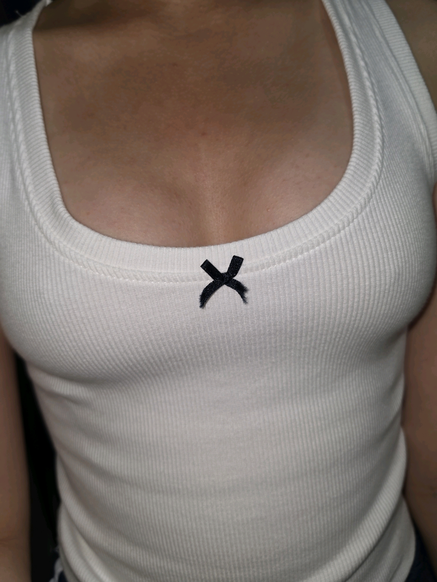 The image shows a close-up view of a person wearing a white ribbed tank top with a small black bow in the center. The person's chest and upper torso are visible. The background is dark and out of focus. There is no discernible location in the city of Phan Thiết, Vietnam.