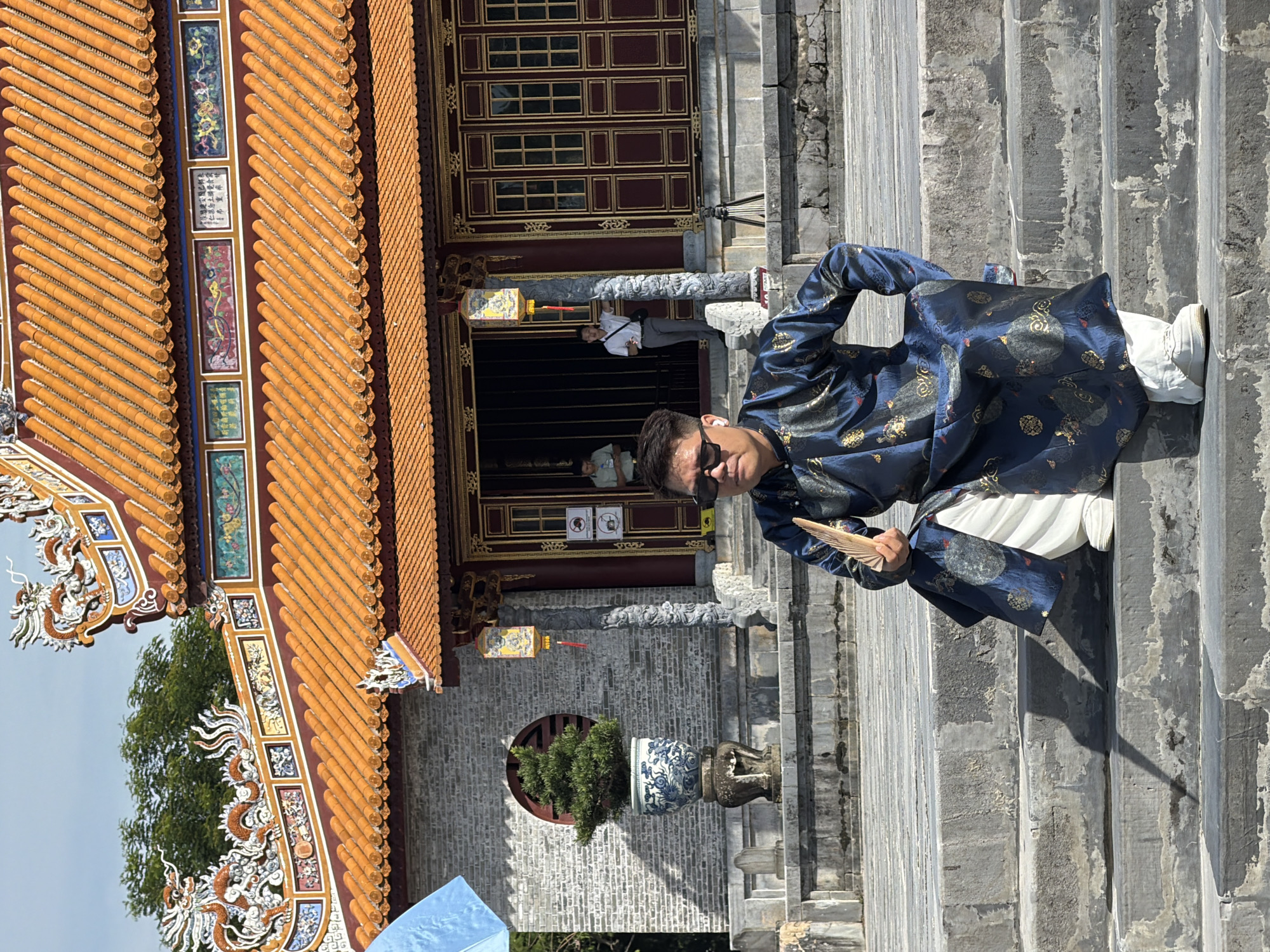 In Phú Xuân Ward, Vietnam, a man dressed in a dark blue robe with gold embellishments sits on stone steps in front of a traditional building. The building features an orange tiled roof with intricate dragon decorations and ornate windows. The man is wearing sunglasses and holding a fan in his right hand. He has white earbuds in his ears. Behind him, two other individuals are visible near the entrance of the building. The environment appears to be a historical site or temple, with stone pillars and a large potted plant with a tree inside situated to the left of the building. A blue umbrella is partially visible in the upper left corner of the frame. The overall scene suggests a cultural or tourist location.