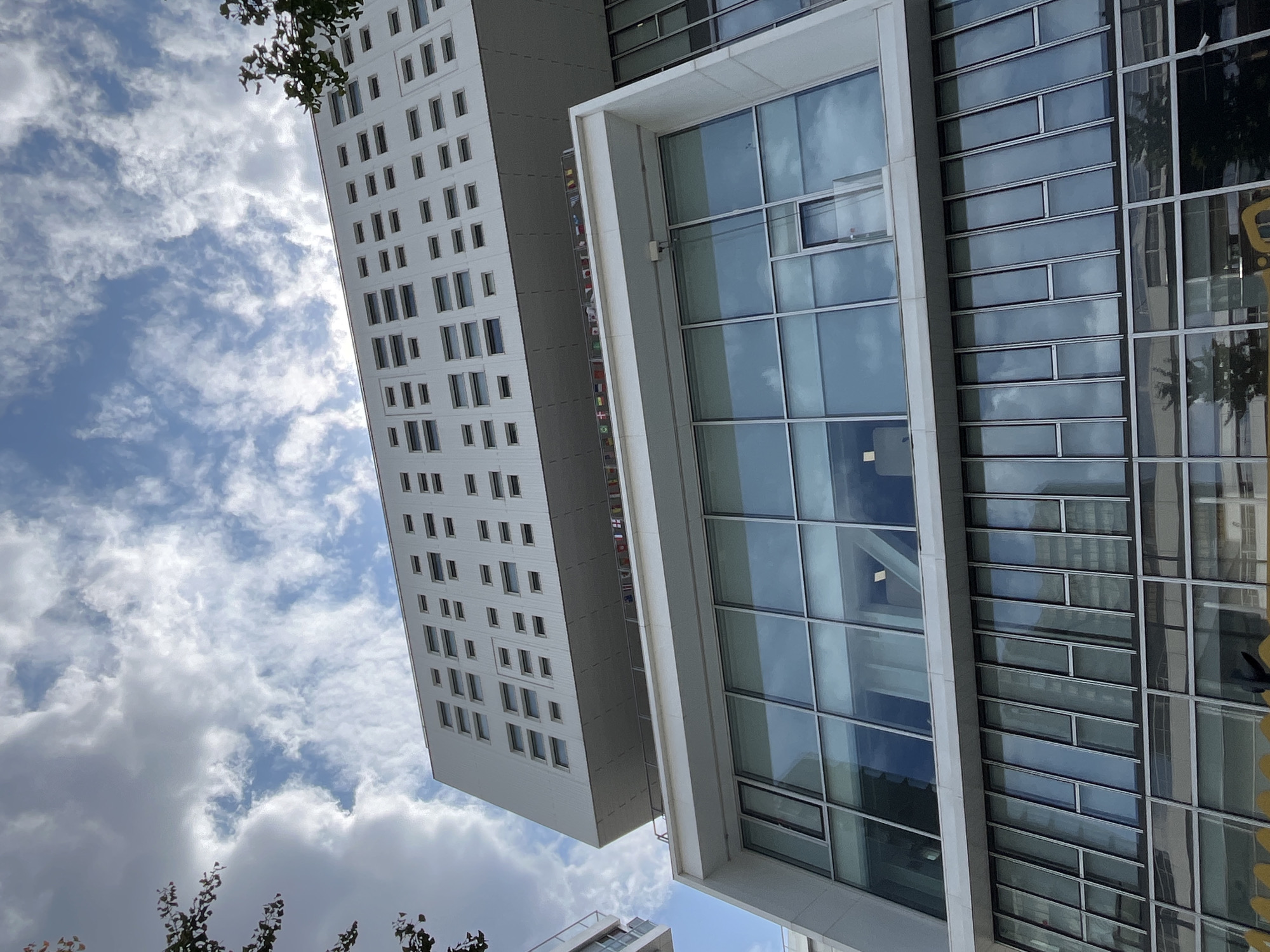 This media file captures an outdoor architectural scene featuring two modern buildings set against a partly cloudy sky. The main subject is a tall, white building with a repetitive pattern of dark, rectangular windows, reminiscent of a grid. Adjacent to this, and partially obscuring it, is another building with a more complex facade. This second building is characterized by large glass panels that reflect the sky and surrounding environment, interspersed with sections of opaque, horizontally oriented rectangular panels.
A prominent architectural element is a white, protruding frame or section that highlights the glass facade of the second building. Along the side of the white building, a vertical strip of small, colorful elements, possibly flags or banners, is visible. The perspective is from below, looking upwards, which emphasizes the height and scale of the structures. A few branches of a leafy tree are visible in the upper left corner, adding a natural element to the urban landscape.
The time of day appears to be daytime, judging by the bright sky and the reflections on the glass. The weather is partly cloudy, with a mix of blue sky and white, fluffy clouds. There is no clear indication of the specific location, nor are there any people or specific activities depicted. The overall mood is one of modern, clean architecture bathed in natural light. No visible text is present in the image.