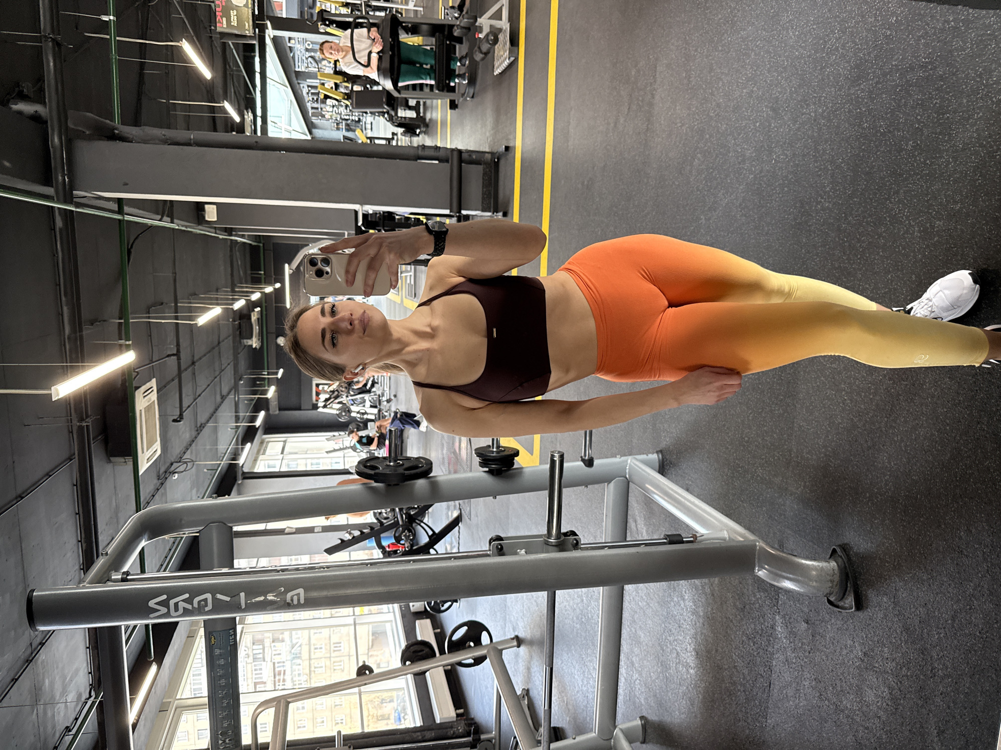 The image shows a woman in a gym, presumably taking a selfie. She is wearing a maroon sports bra and orange-to-yellow gradient leggings, with white sneakers. She is holding her phone in her right hand, with a watch visible on her wrist. The gym is well-equipped, with various weight machines and equipment visible in the background. Light fixtures are visible on the ceiling. There is a window on the side of the machine.
Based on the architectural style visible through the windows, it appears to be Saint Petersburg, Russia. The buildings visible through the windows appear to be typical of the city's architecture.