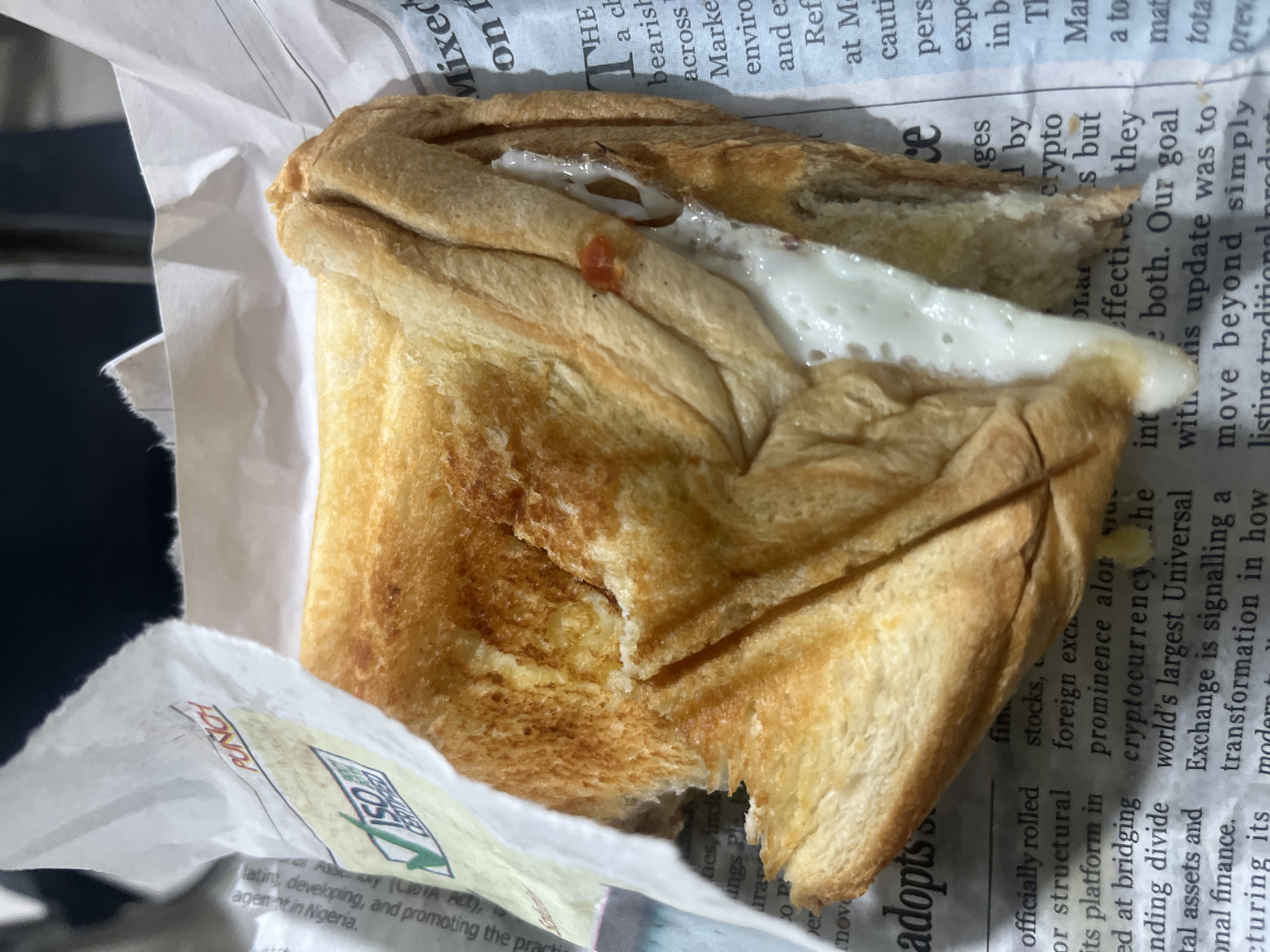 A partly eaten sandwich is wrapped in a newspaper with a "PUNCH" label and visible text mentioning cryptocurrency, stock markets, and Nigeria. The food is likely from Ikeja, Nigeria, based on the newspaper information.
