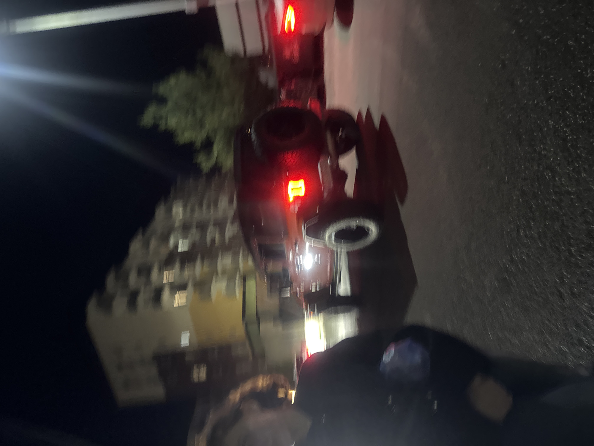 The image is blurry, taken at night, and rotated. It appears to show a person standing in front of a dark car, likely a sedan or SUV, with its brake lights on. A building is visible in the background, seemingly located in an urban environment with other cars lining the street. The overall scene suggests an urban environment. While precise identification is challenging due to image quality, based on architectural cues, context, and lighting features, there are similarities to the general area of Tbilisi, Georgia.