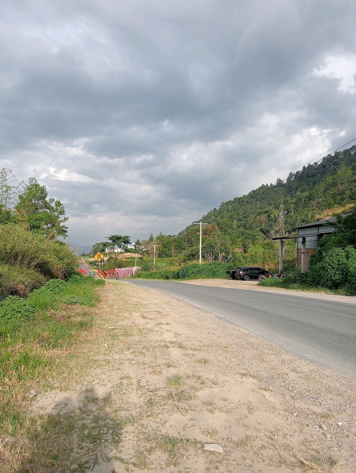 The image depicts a rural road scene in Hinalangbagasan, Indonesia, under a cloudy sky. On the left side of the road, there is lush green vegetation with tall grass and bamboo-like plants lining a ditch. A red and yellow bridge with decorative banners is visible, and beyond it, a yellow traffic sign indicating a curve to the right. A motorcycle is parked near the bridge. The road itself is paved, with a dirt and gravel shoulder. On the right side of the road, a steep, tree-covered hill dominates the background. A dark-colored SUV is parked near a simple, open-air structure with a corrugated metal roof. A power pole stands near the SUV. The overall atmosphere suggests a quiet, natural setting.