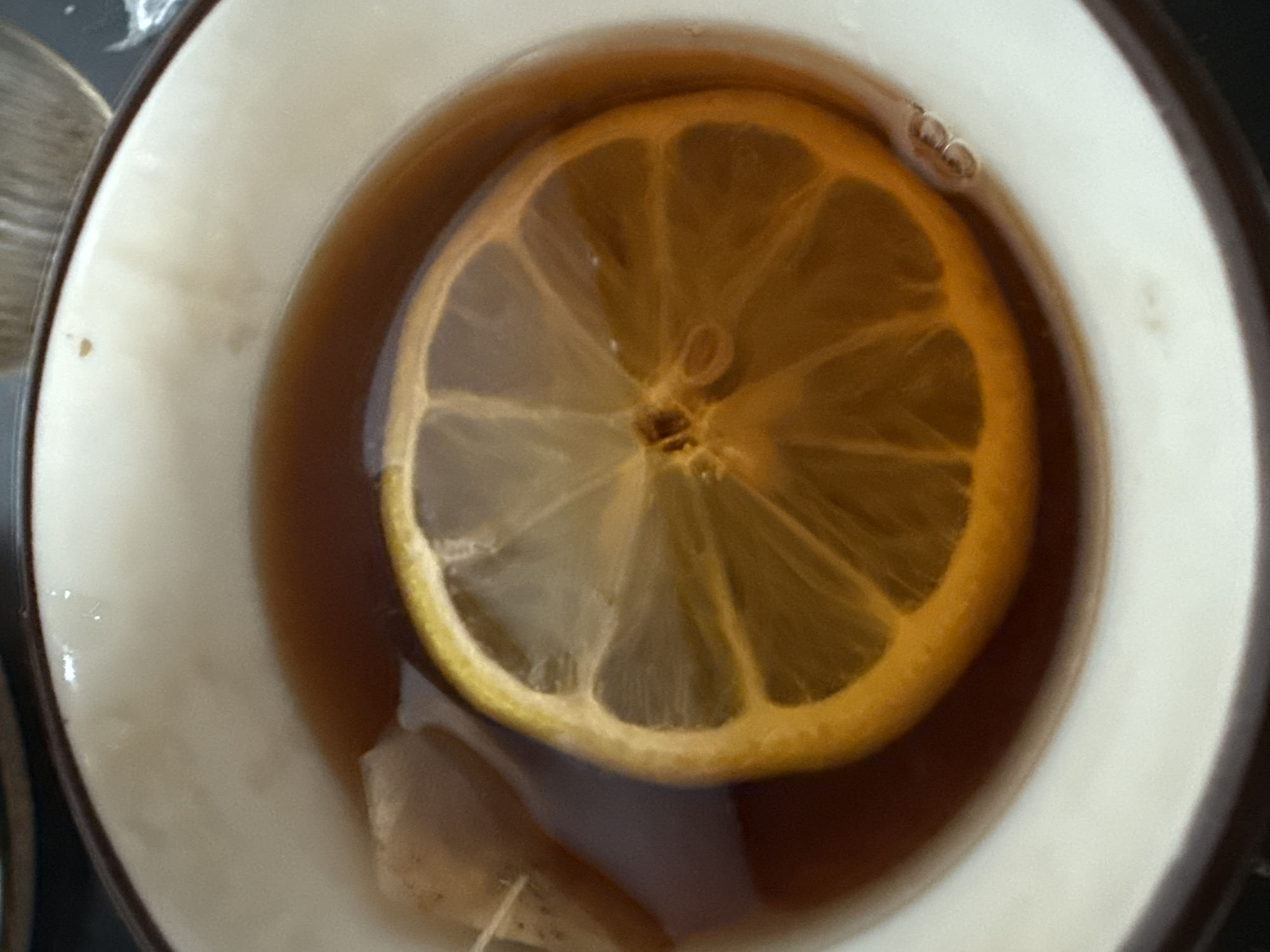 A close-up, high-angle shot shows a white teacup filled with dark brown liquid, likely tea. A lemon slice occupies the center of the cup, partially submerged in the tea. At the bottom of the cup, a tea bag tag is visible, suggesting a tea bag is steeping in the beverage. Several small bubbles can be seen near the edge of the lemon slice. Part of another cup appears to be in the view to the left of the cup in focus. No people, subjects, objects, actions, interactions, and no indication of the city of Vidnoye, Russia are depicted in the image.