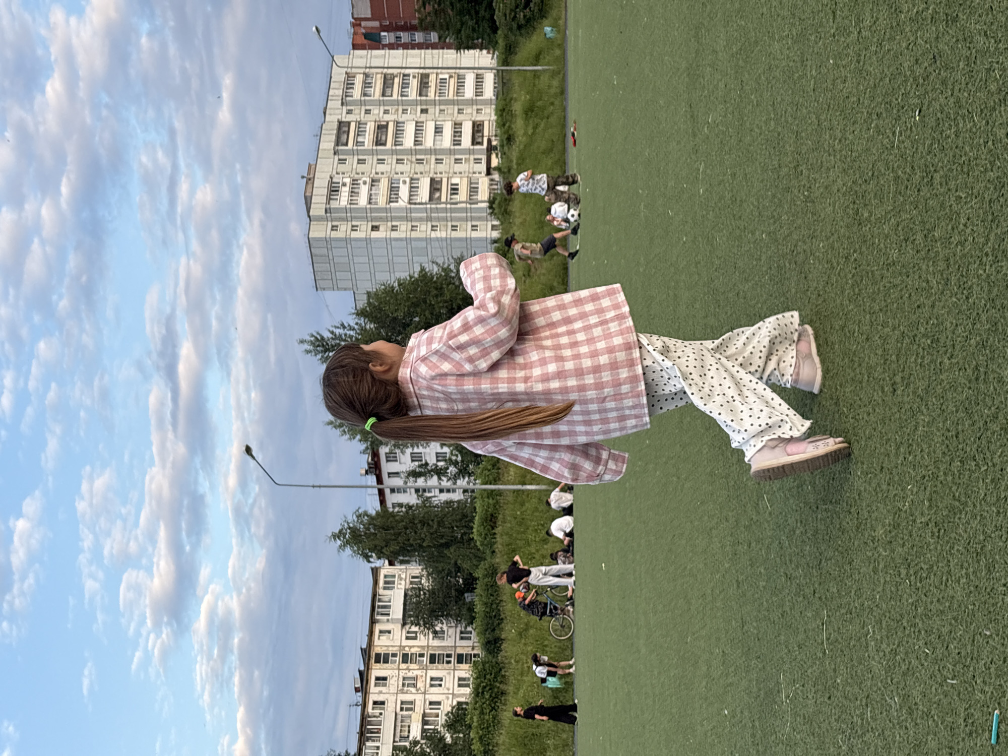 A young girl wearing a pink and white checkered shirt and polka dot pants stands on a green artificial turf field. Her hair is tied back with a green elastic band. In the background, several people are gathered on the field, some appearing to be playing with a soccer ball. A bicycle is also visible. Behind the field, there are trees and tall apartment buildings. The sky is a pale blue with scattered white clouds. The lighting suggests it is daytime, possibly late afternoon or early evening. The setting appears to be a residential area in Chaikovsky, Russia, with a sports field adjacent to apartment complexes. There is no visible text in the image.