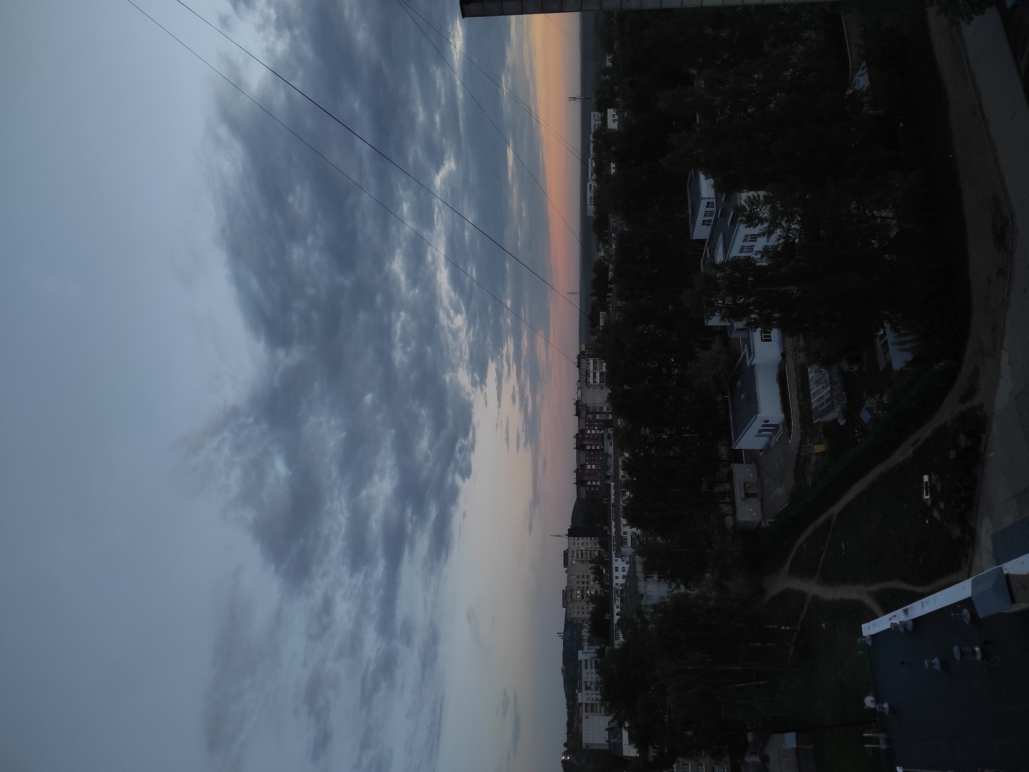 The image presents a cityscape at dusk, likely captured from a high vantage point. The sky, dominating the left portion, transitions from a pale blue-gray to darker, stormy clouds. Several power lines stretch across the upper-left quadrant. A thin band of orange and pink hues separates the sky from the city below.
The city itself appears as a mix of residential buildings and trees. Some buildings are taller and denser, typical of urban centers. Green areas suggest parks or tree-lined streets. The right side of the image shows buildings closer to the vantage point, with more detailed views of rooftops and possibly paths.
Given the prompt information, it is suggested that this is a landscape shot in the town of Chaikovsky, Russia.