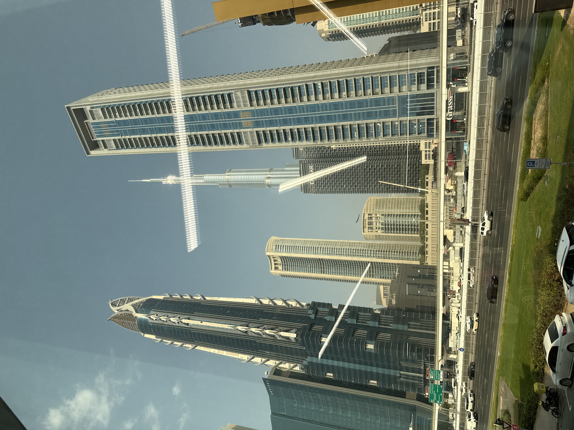 This is an eye-level, medium shot capturing a cityscape of Dubai, United Arab Emirates, dominated by modern skyscrapers. The Burj Khalifa, the world's tallest building, is prominently visible in the background, its distinctive spire reaching towards the clear blue sky. In the foreground, a multi-lane highway is bustling with traffic, including sedans, SUVs, and delivery vehicles. The highway is elevated, with overpasses and infrastructure supporting the flow of vehicles. Surrounding the highway are well-maintained green spaces and sidewalks. The image is taken through a window, as evidenced by reflections of light and a white, horizontal streak across the frame. The architecture suggests the financial district of Dubai, likely near Sheikh Zayed Road, given the density of skyscrapers and the presence of the Burj Khalifa. There are no discernible people or animals in the frame, and the focus is on the urban infrastructure and impressive architectural achievements.