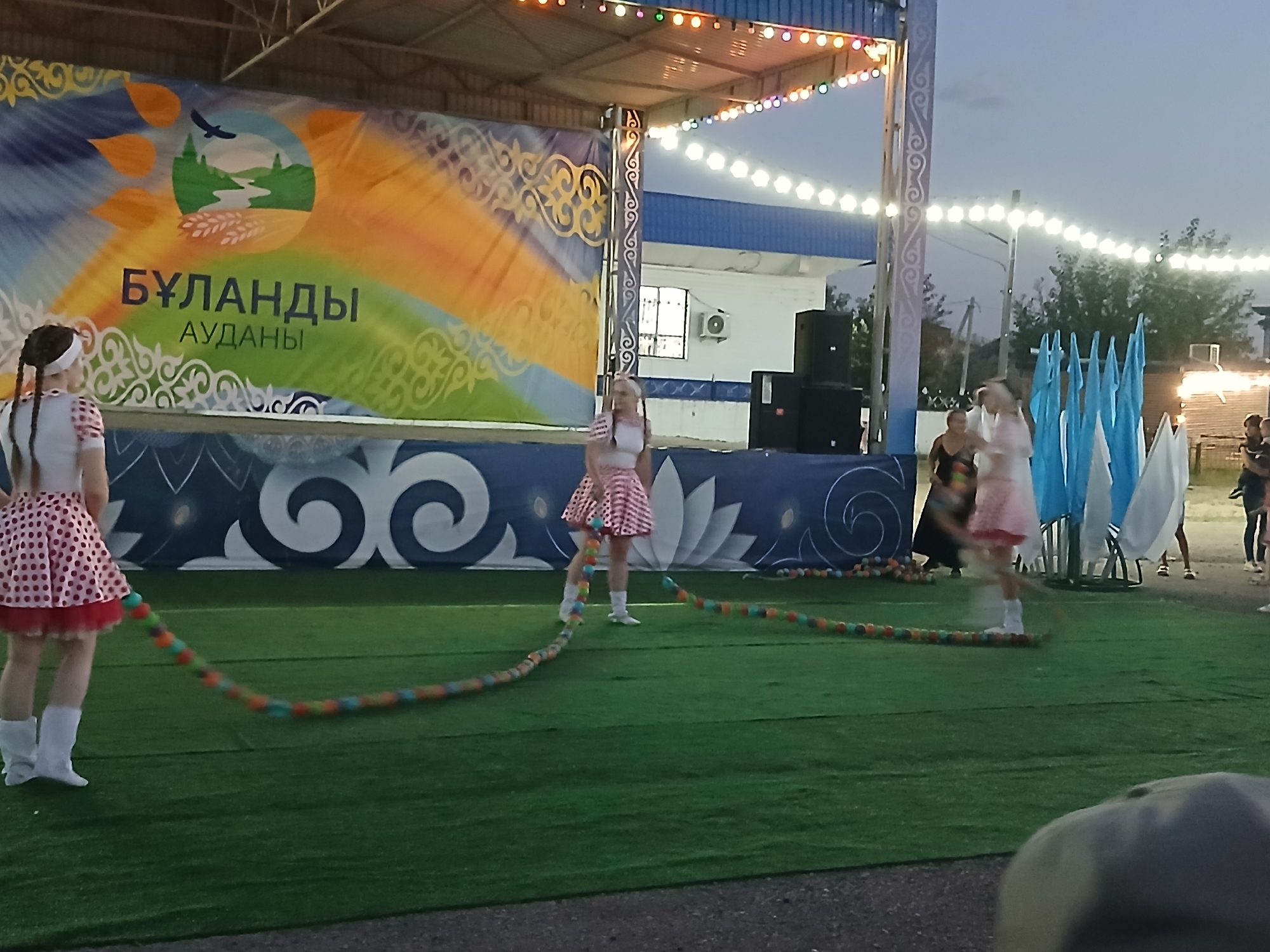 In Makinsk, Kazakhstan, an outdoor evening event features a stage with a large banner displaying the text "БҰЛАНДЫ АУДАНЫ" in Kazakh, beneath an illustration of a sun, landscape, and agricultural imagery. The stage is illuminated by string lights, and speakers are visible on either side. On the artificial turf in front of the stage, three young girls are participating in a jump rope activity with a colorful rope made of large beads. The girl on the far left wears a red and white polka dot dress and white boots, while the girl in the center has a similar dress and white socks. The girl on the right is partially obscured but appears to be wearing a pink dress. In the background, several adults are present, and a series of light blue and white flags are displayed. The overall environment suggests a community gathering or festival.