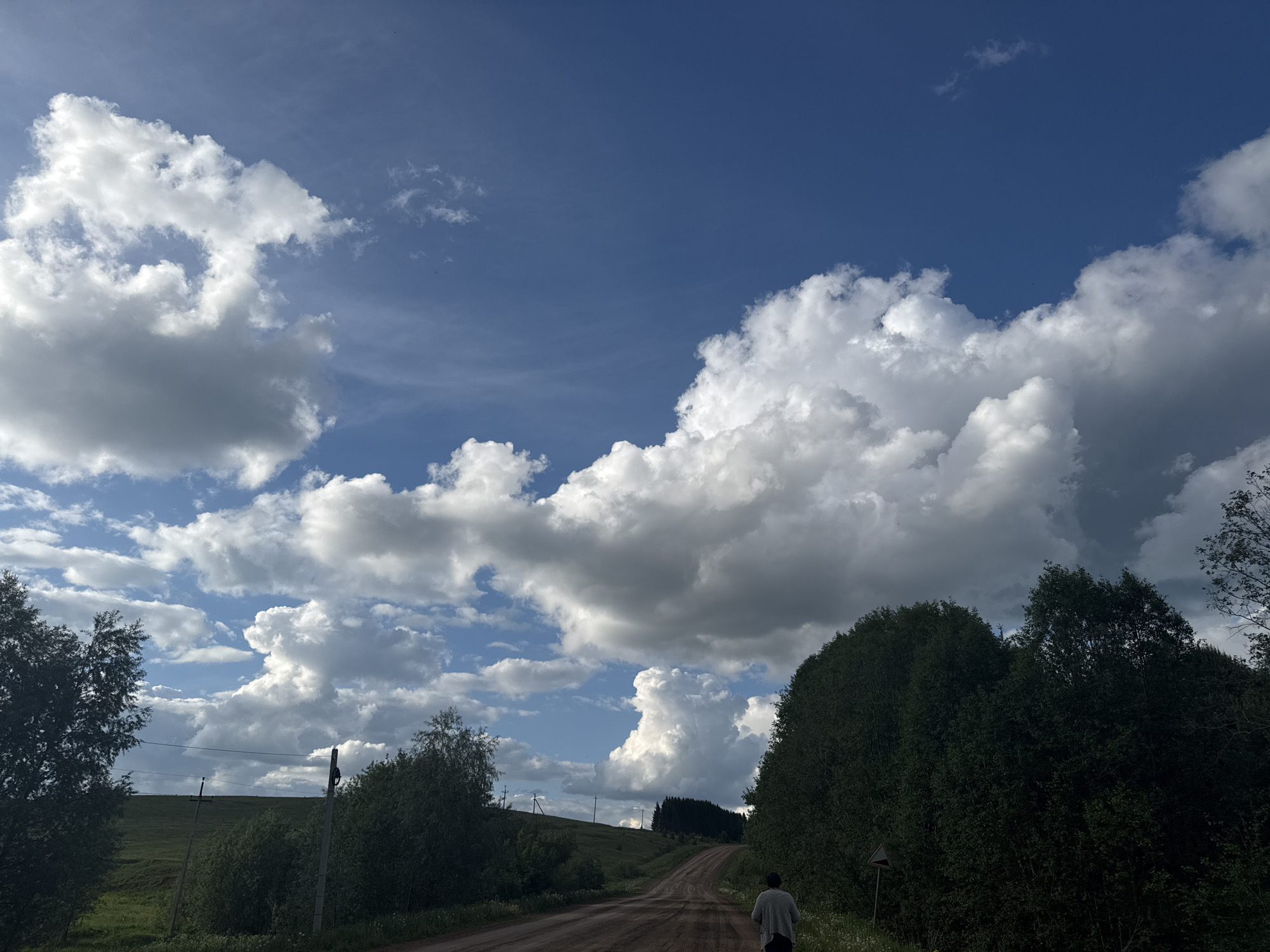 The image depicts a rural scene, likely in the vicinity of Batmany, Russia. A dirt road extends into the distance, leading uphill. The foreground is bordered by green foliage and trees on either side. A single person, facing away from the camera, is walking up the road. The sky is predominantly blue, filled with large, puffy cumulus clouds. In the background, rolling hills and more trees are visible.