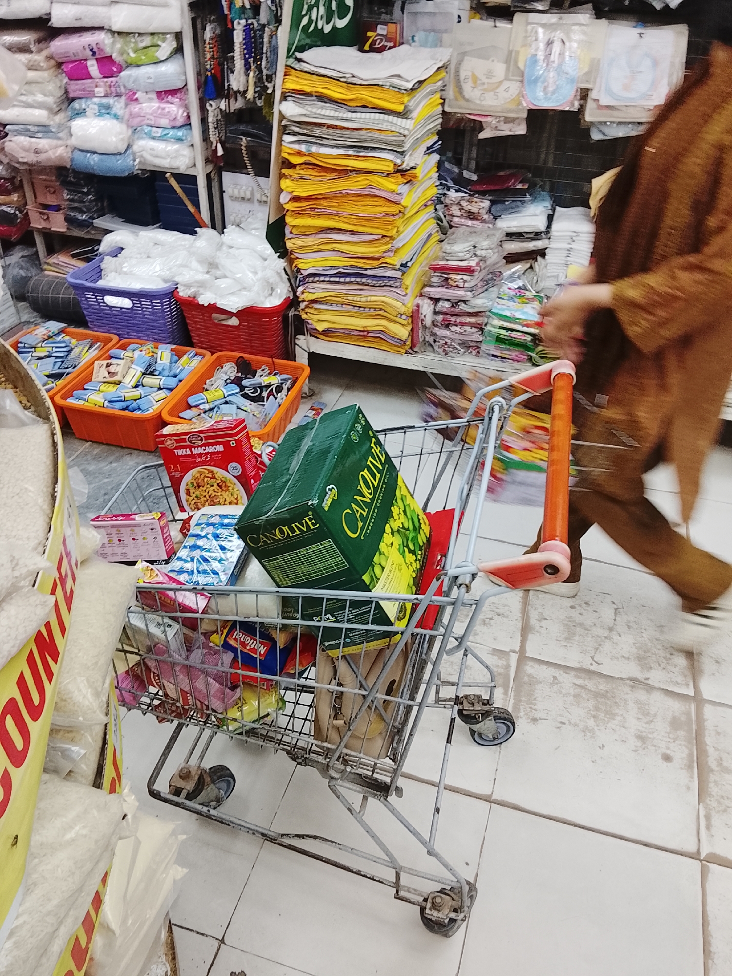 The image shows a shopping cart in a store. The cart contains items such as a box of Canolive oil, a box of Tikka Macaroni, and various snack foods. Shelves in the background hold stacked textiles, packaged goods, and baskets filled with merchandise. A person wearing a brown outfit walks by the cart. The scene appears to be a retail establishment in Gulgasht Colony, Pakistan, based on the product packaging.