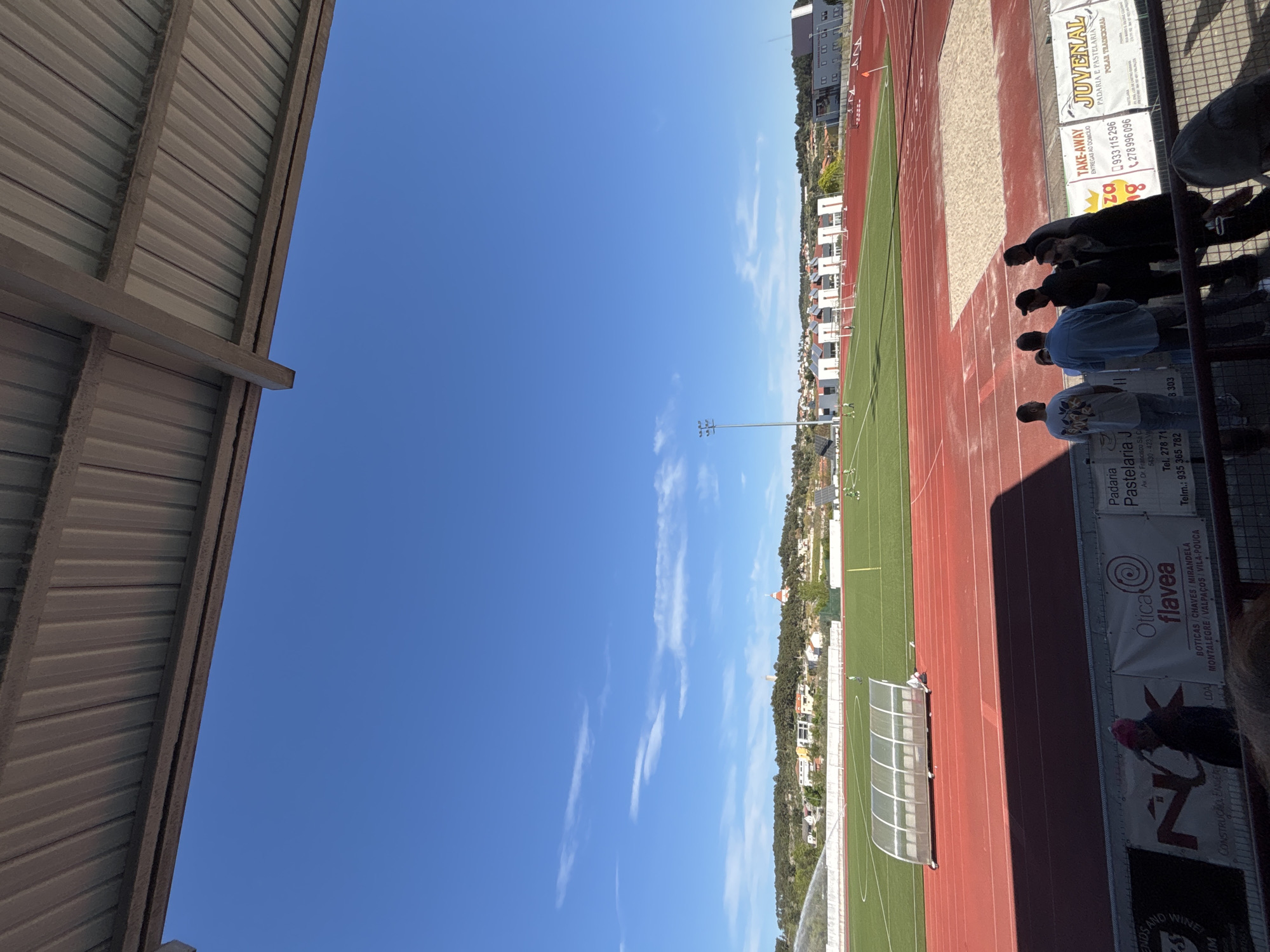 The image depicts a track and field event viewed from stadium seating. On the right side of the frame, several people are visible, with some facing away and others obscured. Signage is attached to the wall with the name “Juvenal Padaria E Pastelaria, Polar Tradicional.” Other signage displays contact information and business details. The track area consists of red running lanes and a green field, and an awning-like structure is at the corner of the track. In the background, buildings and a townscape are visible. Above the townscape is a clear blue sky with sparse clouds. The image seems to be taken in Valpaços, Portugal, based on the business signs visible. A metal stadium overhang is on the left side of the frame.
