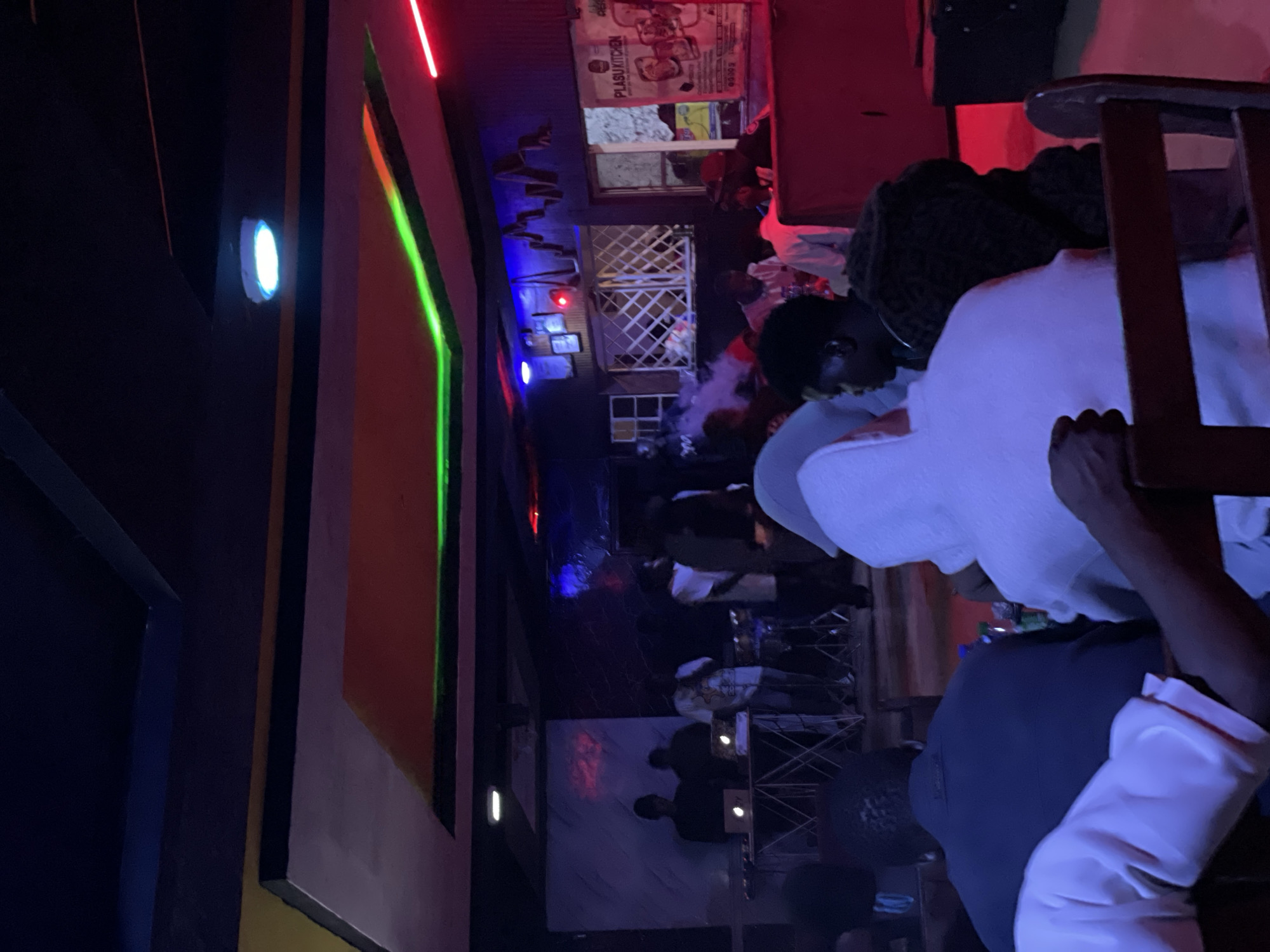 The image depicts an indoor gathering at a venue identified as "Plasu Kitchen" in Durumi, Nigeria. The atmosphere is dimly lit with vibrant neon lighting, including green and red strips along the ceiling and a blue spotlight. In the background, a DJ setup with laptops and drums is visible, suggesting a musical performance or event. Several individuals are present, some standing and some seated, engaged in conversation or observing the scene. The patrons are casually dressed, with some wearing hoodies. The overall impression is that of a lively social gathering or entertainment venue.