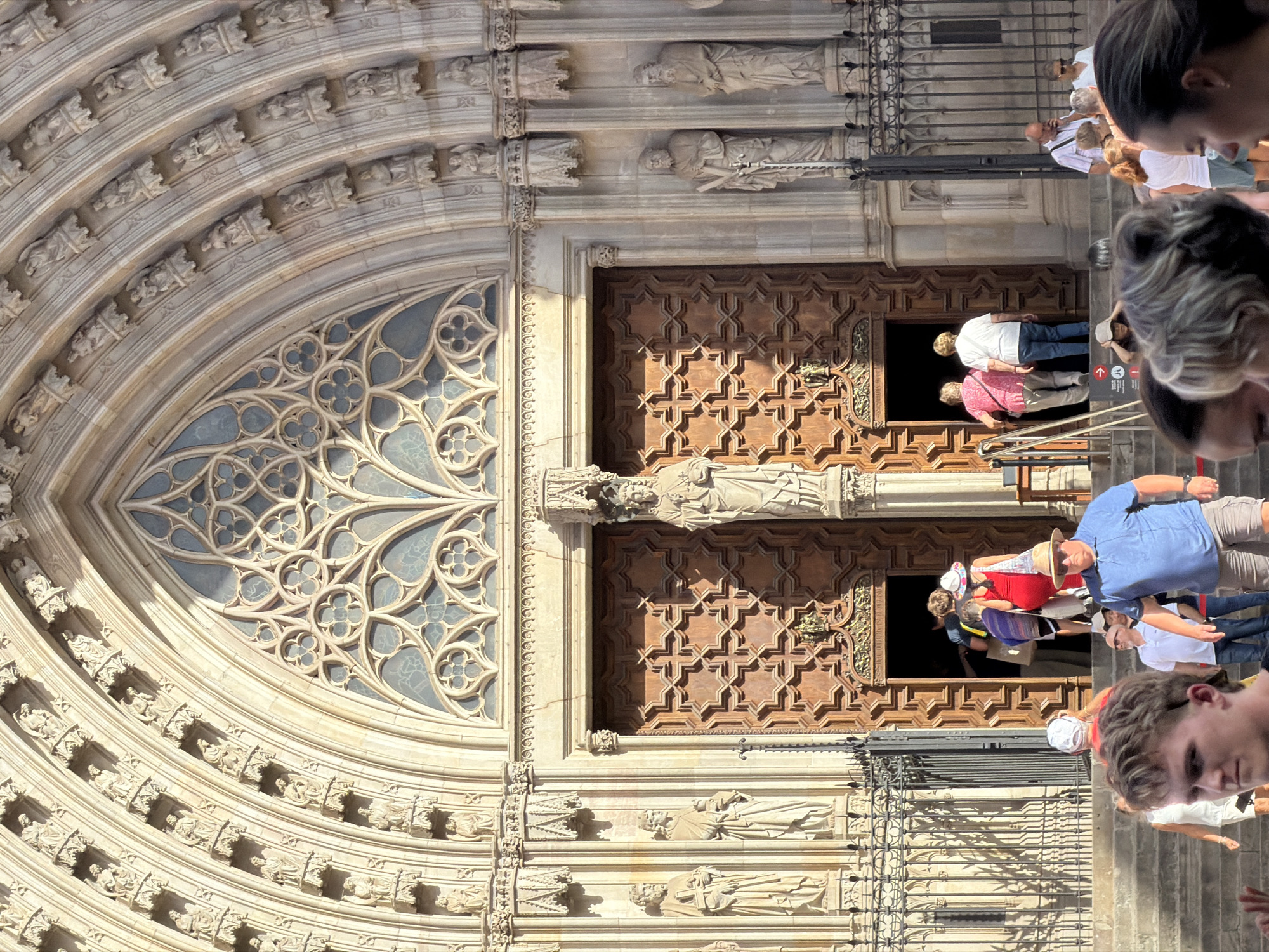 The image depicts the ornate facade of the Barcelona Cathedral in Barcelona, Spain, bathed in sunlight. The cathedral's entrance features grand wooden doors with intricate geometric patterns and a large rose window above, adorned with Gothic tracery. Flanking the entrance are stone statues of figures in robes, and the arched entryway is decorated with numerous small niches containing more sculpted figures.
In front of the cathedral, a group of tourists are seen entering and exiting the building. Some are dressed casually in t-shirts, shorts, and hats, suggesting warm weather. One man in the foreground, with blonde hair and a mullet, is looking towards the camera, possibly posing or captured candidly. People are walking up and down the wide stone steps leading to the entrance. A black wrought-iron fence runs along the side of the steps, and shadows from the fence are cast on the stone. The scene captures a typical day at a popular tourist attraction, with a mix of architectural grandeur and the bustle of visitors.