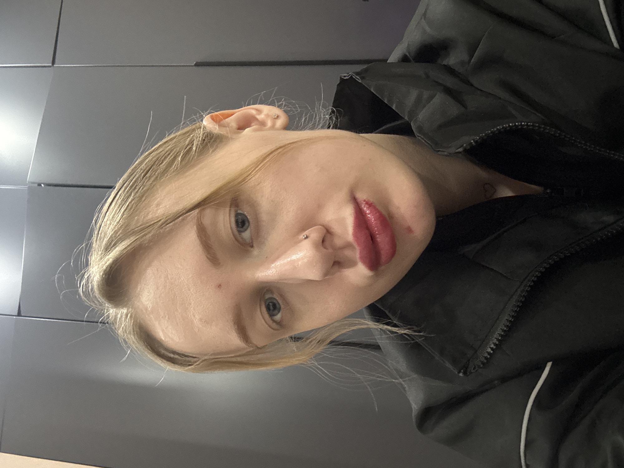 A close-up, slightly angled selfie shows a fair-skinned blonde woman wearing a black jacket with a white stripe visible near the zipper. She has light blue eyes, a piercing in her left nostril, and her lips are prominently coated in red lipstick. A small tattoo, potentially a heart, is visible on her chest above the jacket. The background is a plain, dark gray wall composed of rectangular panels. The image provides no contextual information about the location being in Novoaleksandrovka, Russia.