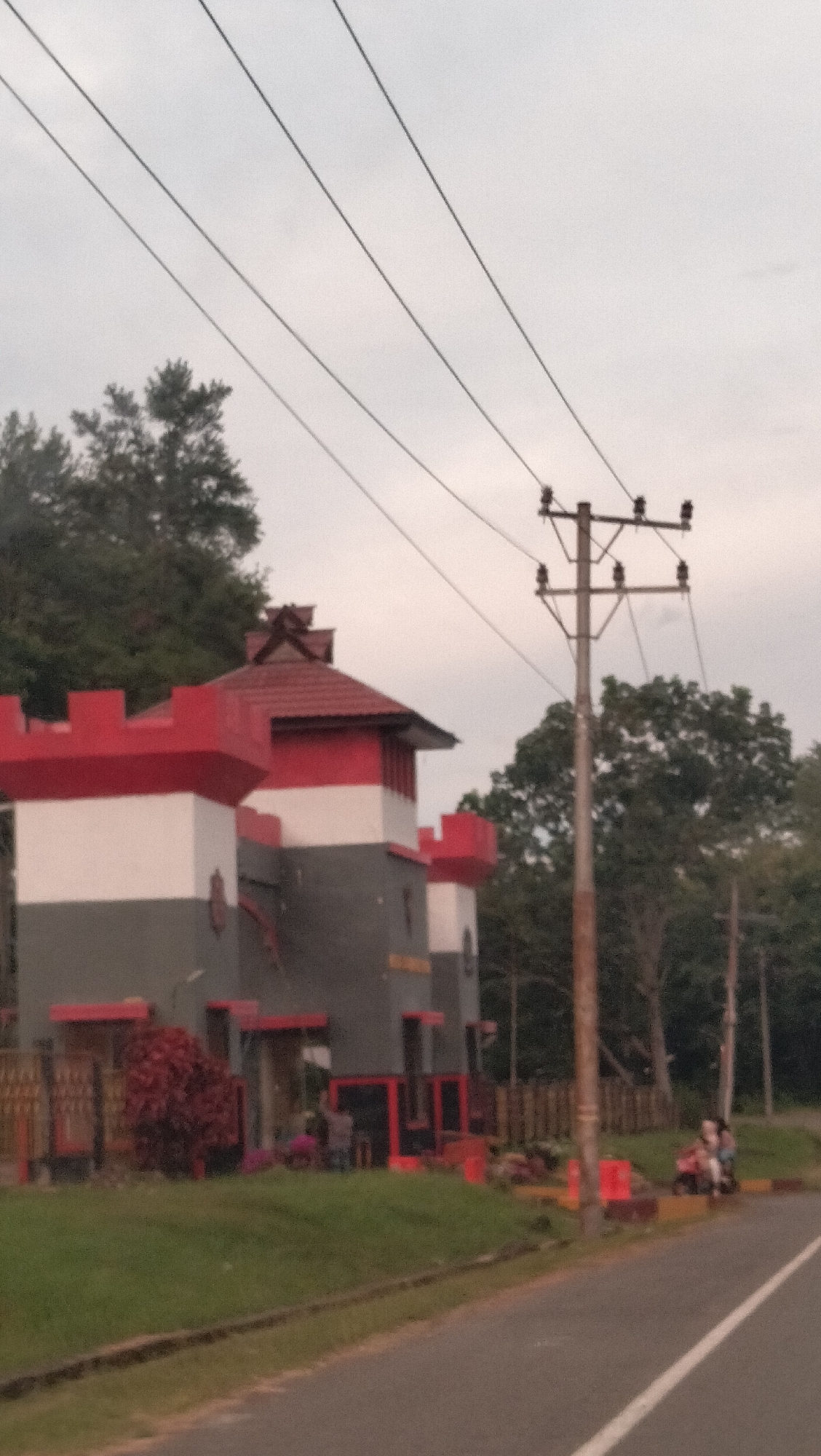 This image from Lamtamot, Indonesia, showcases a distinctive castle-like building alongside a road under an overcast sky. The building features a unique design with horizontal sections of red, white, and grey, topped by a red tiled roof displaying traditional Indonesian architectural motifs. Red-framed windows or entrances and small red fences are visible at its base.
In front of the building, there is green grass and a bush with reddish foliage. Several indistinct human figures can be seen near the building, with at least two individuals standing near a red structure. To the right of the building, a wooden utility pole with multiple power lines extends upwards, contrasting against the light grey sky. Dense green trees form the backdrop behind the building and the utility pole.
In the foreground, an asphalt road with a white median line runs along the bottom and right side of the frame. On the far right, two people are seated on a motorbike by the roadside. The soft, diffuse lighting suggests an overcast day, possibly in the late afternoon or early morning. Indistinct white text is faintly visible on the grey section of the building.